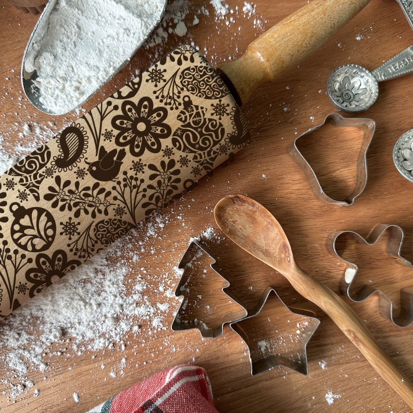 Wooden rolling pin with decorative patterns, flour, metal cookie cutters, and wooden spoon on a wooden surface.