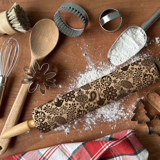 Baking tools including a rolling pin with embossed designs, wooden spoon, whisk, and cookie cutters on a wooden surface.