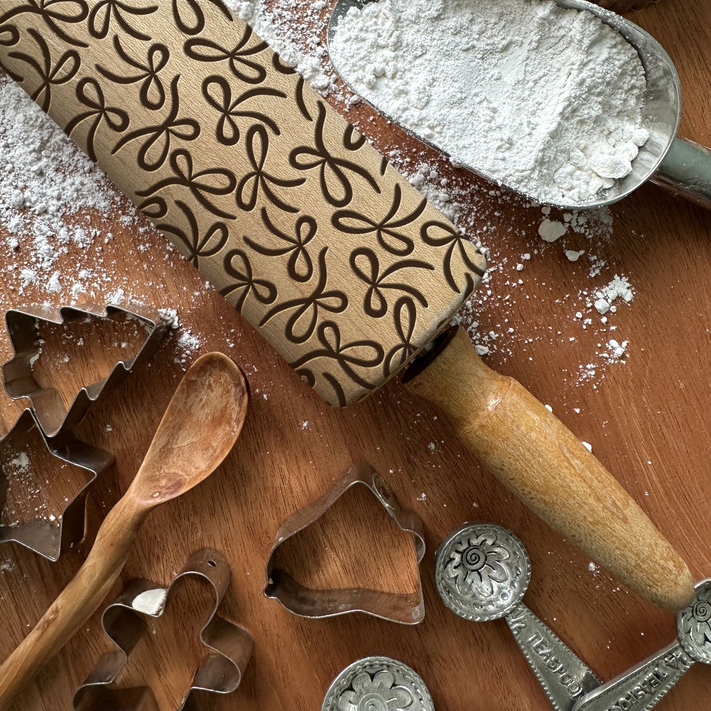 Baking tools including a rolling pin with a pattern, cookie cutters, and a wooden spoon on a wooden surface.