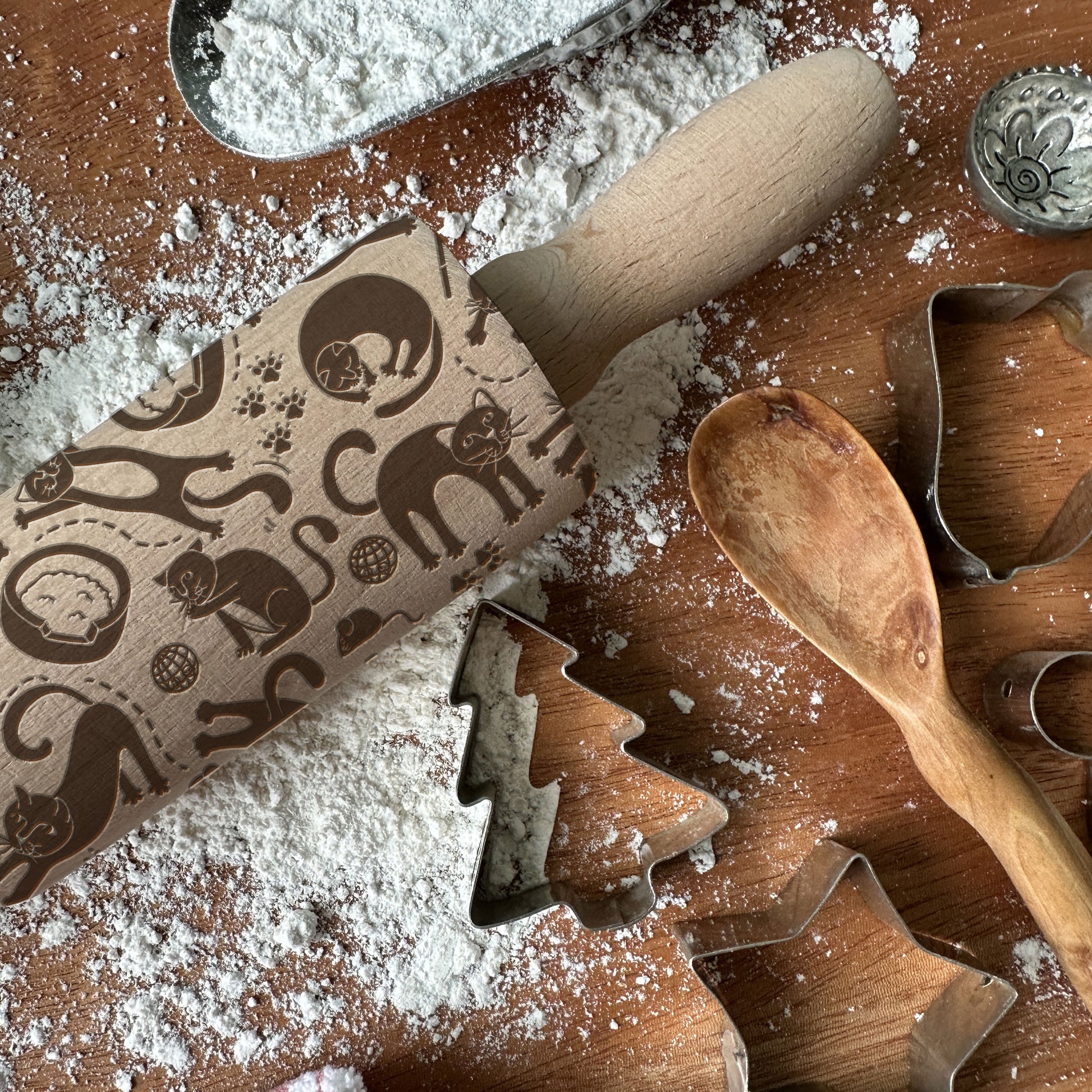 Baking tools including a rolling pin with cat patterns, wooden spoon, and metal cookie cutters on a flour-dusted surface.