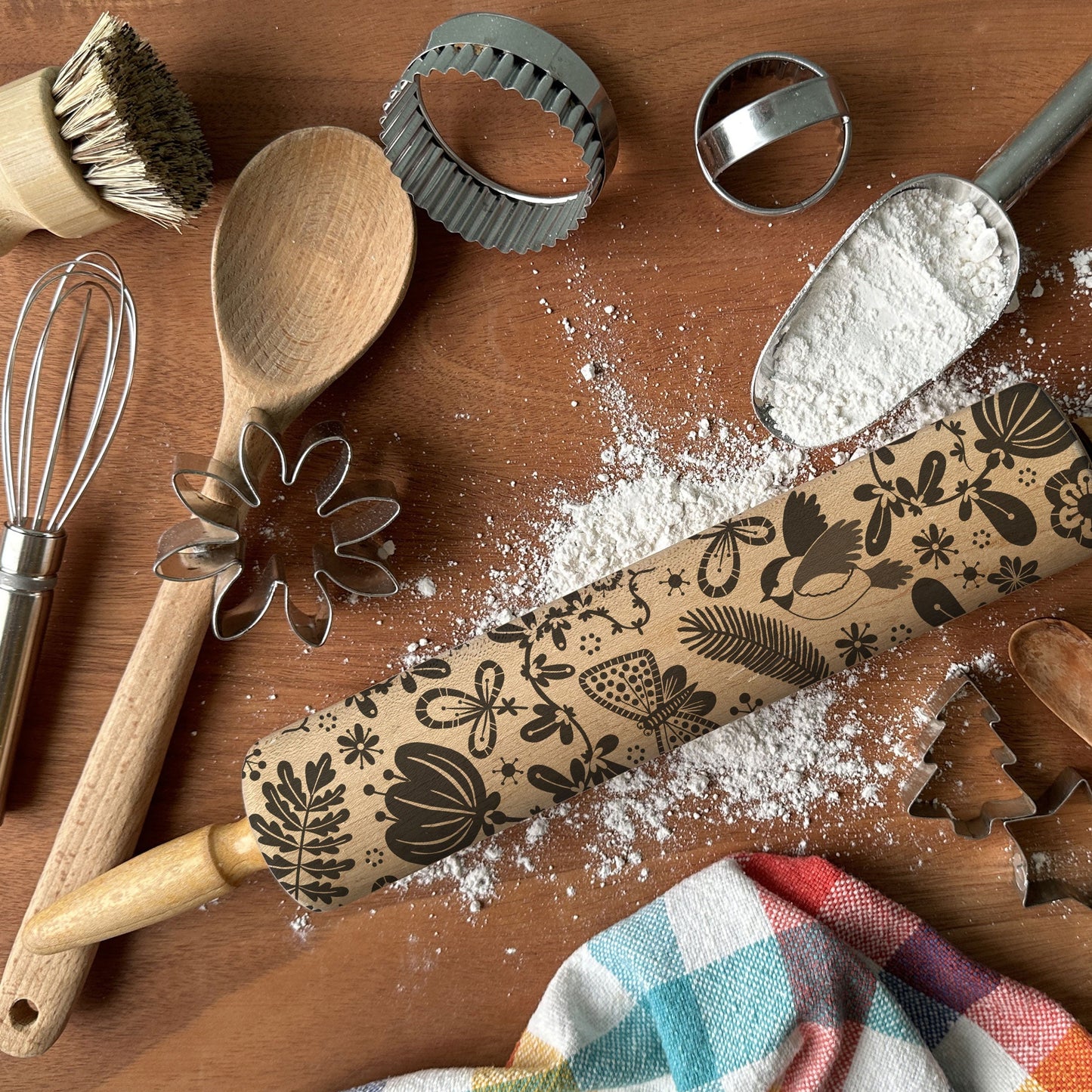 Baking tools including a rolling pin with floral patterns, wooden spoon, whisk, and cookie cutters on a wooden surface.
