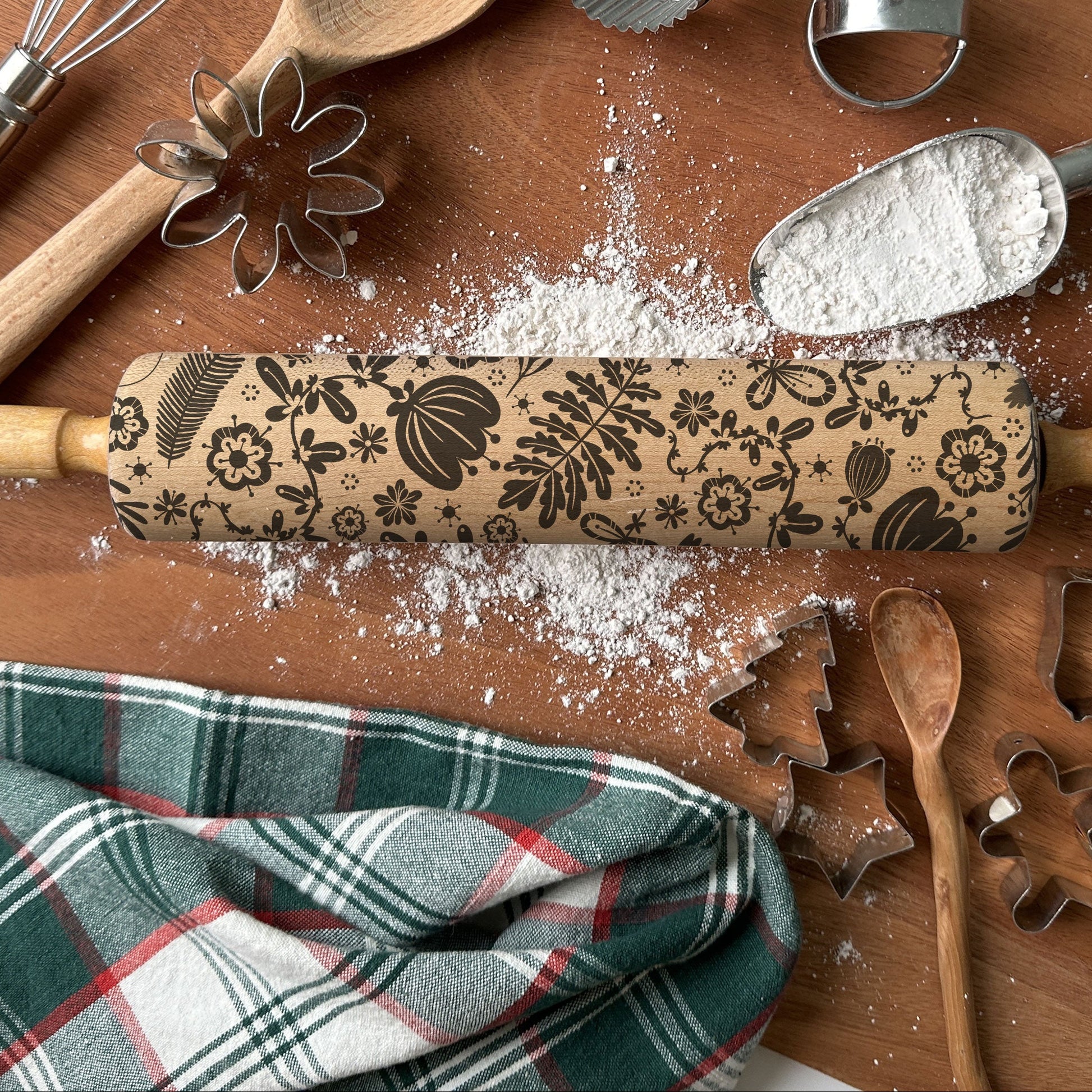 Baking tools including a rolling pin with floral patterns, wooden spoon, whisk, and plaid towel on a wooden surface.