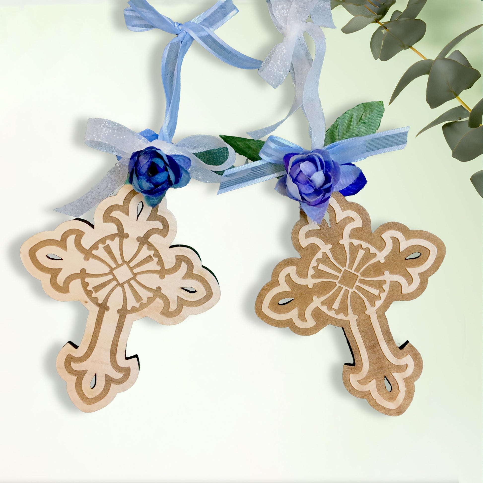 Two decorative cross ornaments with blue ribbons and flowers on a light green background