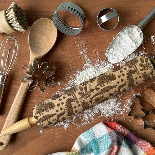 Baking tools including a rolling pin with embossed designs, wooden spoon, whisk, and cookie cutters on a wooden surface.
