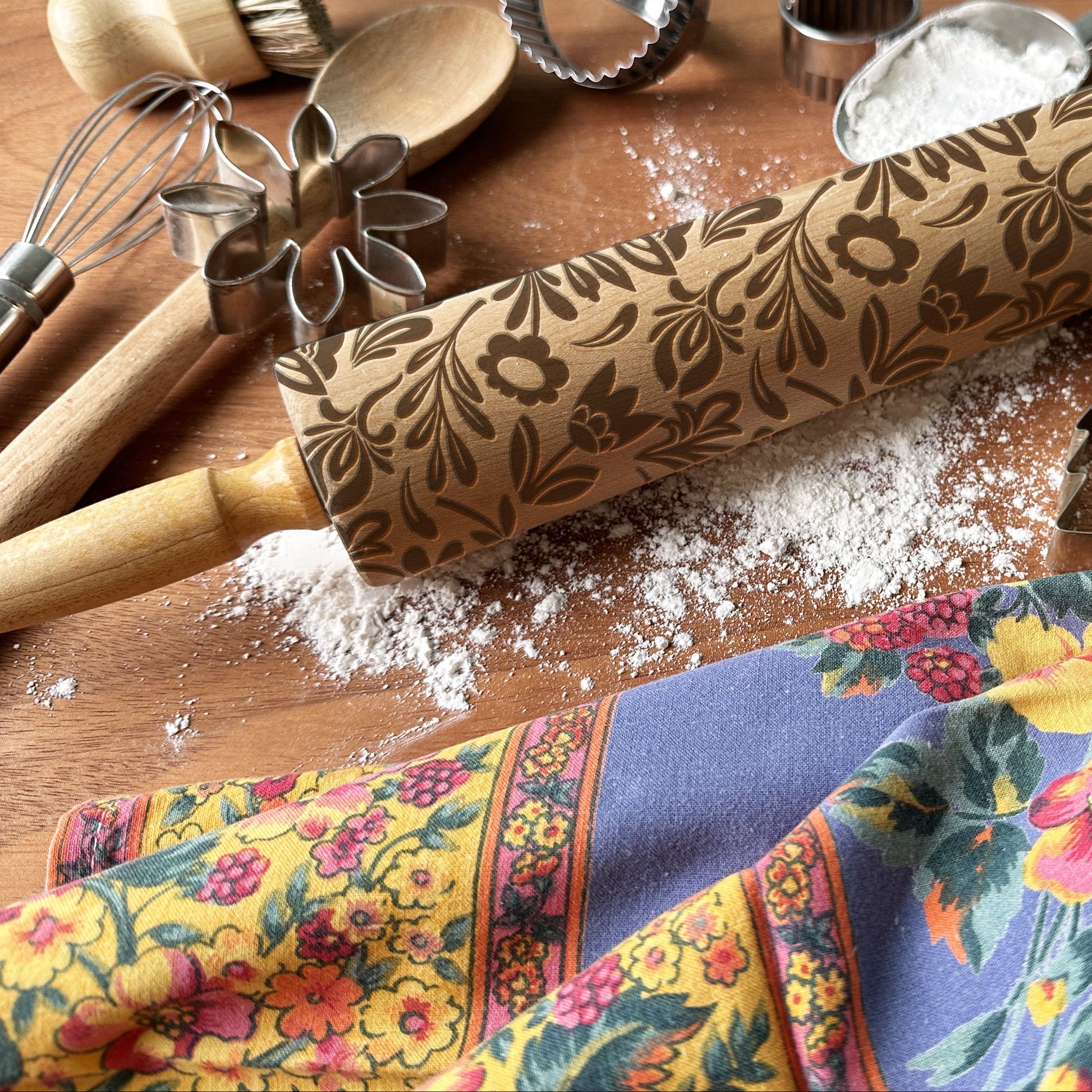 Decorative rolling pin with floral pattern on a wooden surface with kitchen utensils and a colorful towel.