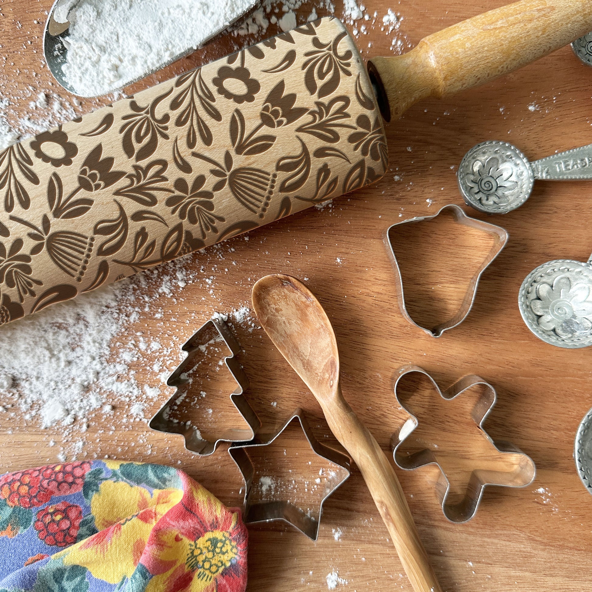 Baking tools including a patterned rolling pin, cookie cutters, and measuring spoons on a wooden surface with flour.