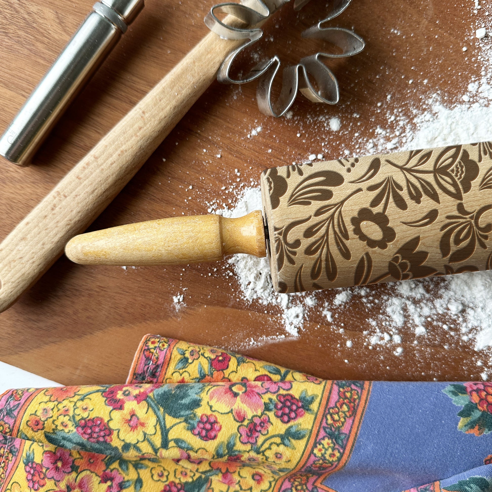 Decorative rolling pin with floral pattern on a wooden surface with flour and a colorful kitchen towel.