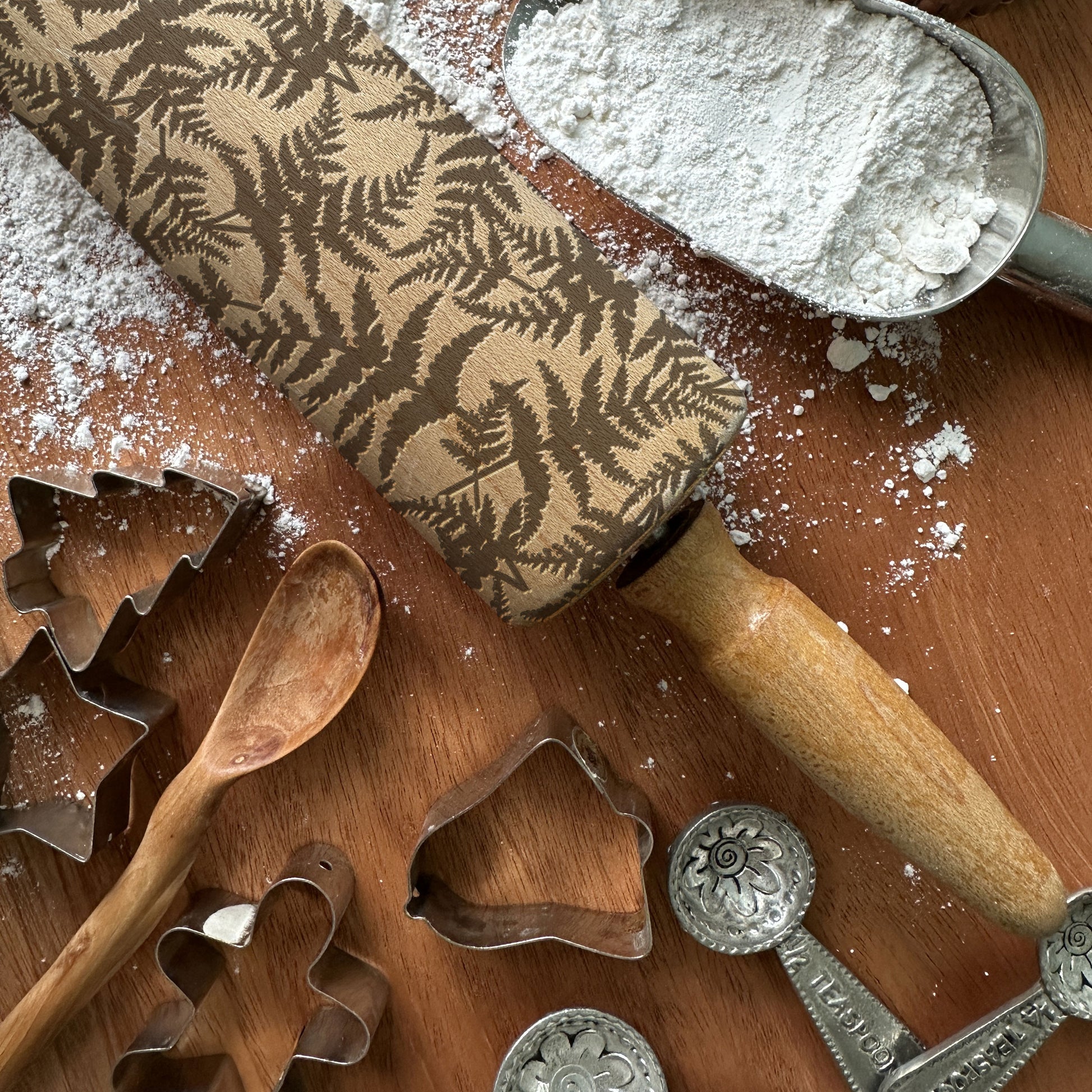 Baking tools including a patterned rolling pin, flour, wooden spoon, and metal cookie cutters on a wooden surface.