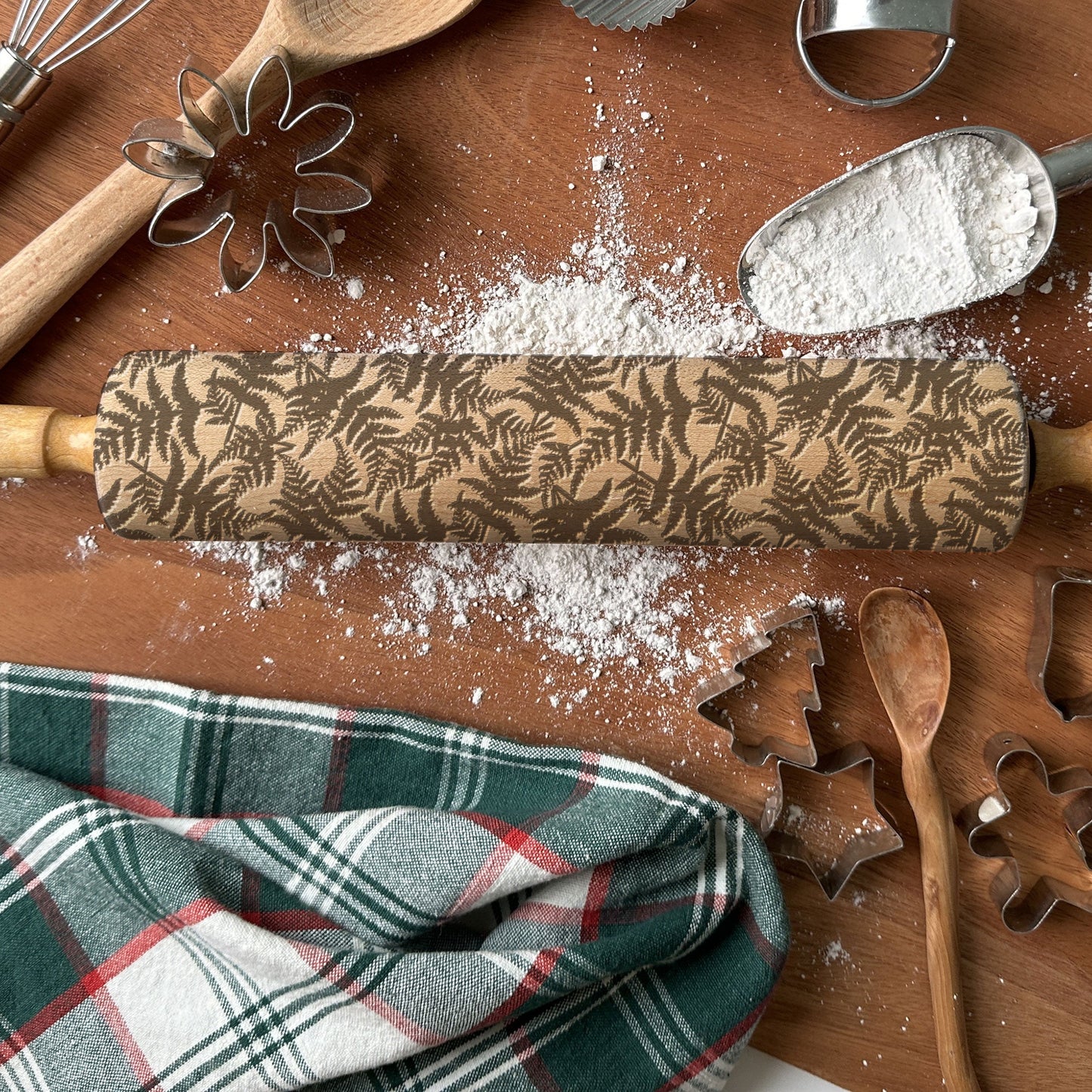 Wooden rolling pin with leaf pattern on a wooden surface with flour, cookie cutters, and a plaid cloth.