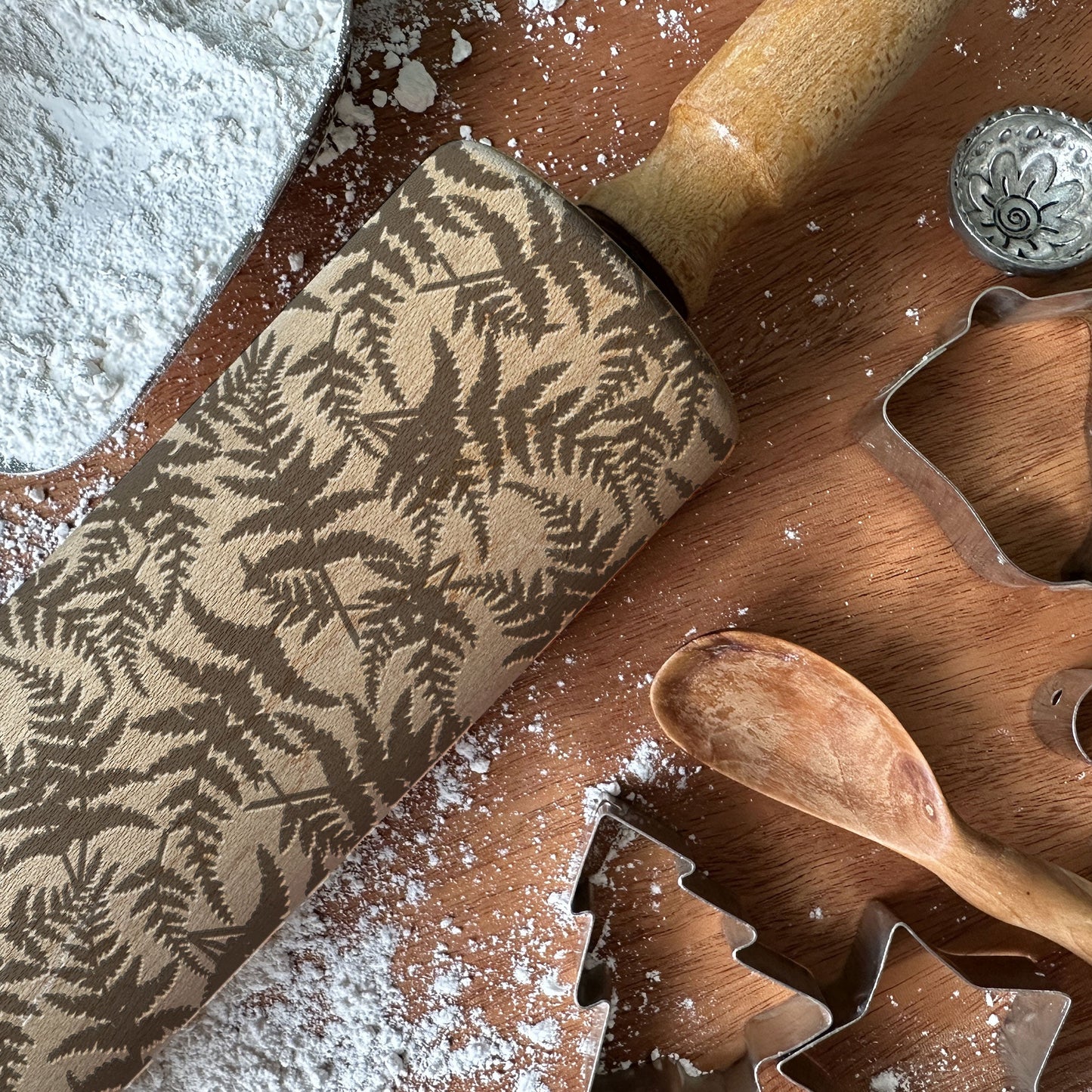 Wooden rolling pin with leaf pattern and cookie cutters on a wooden surface with flour.
