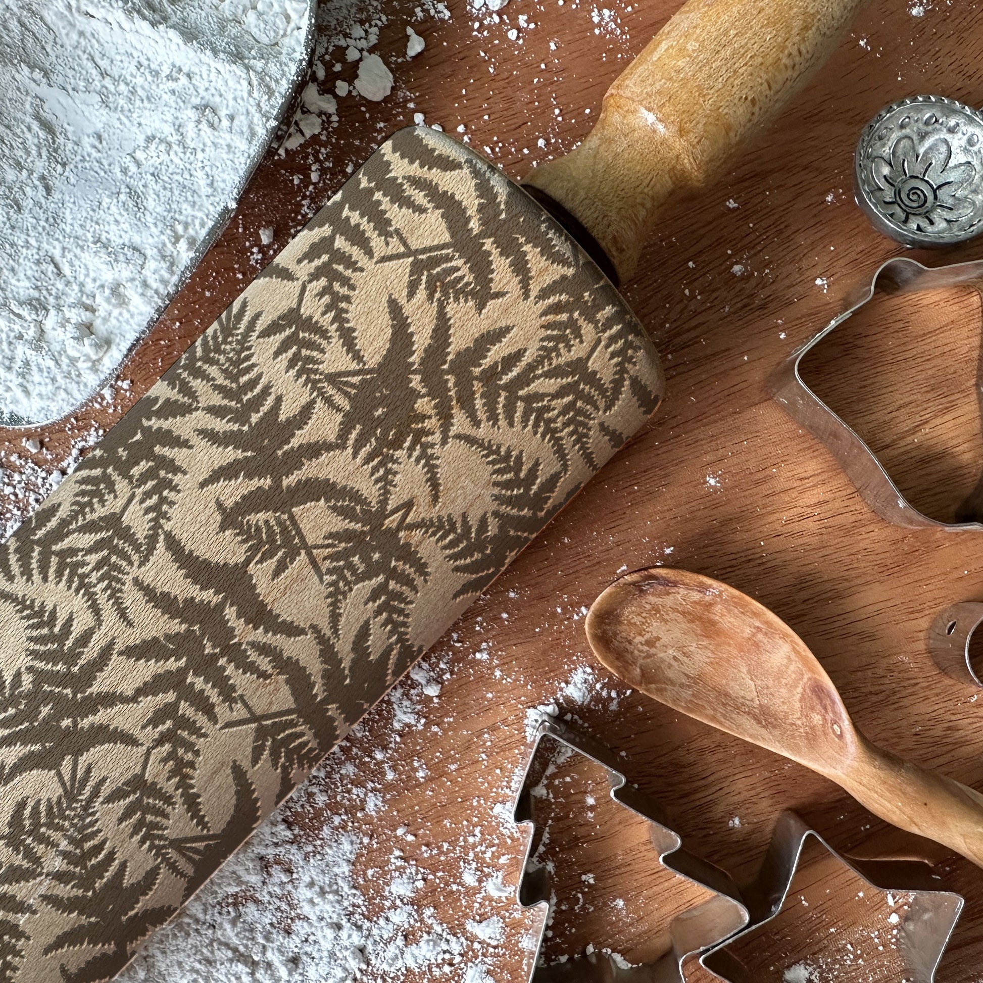 Wooden rolling pin with leaf pattern and cookie cutters on a wooden surface with flour.
