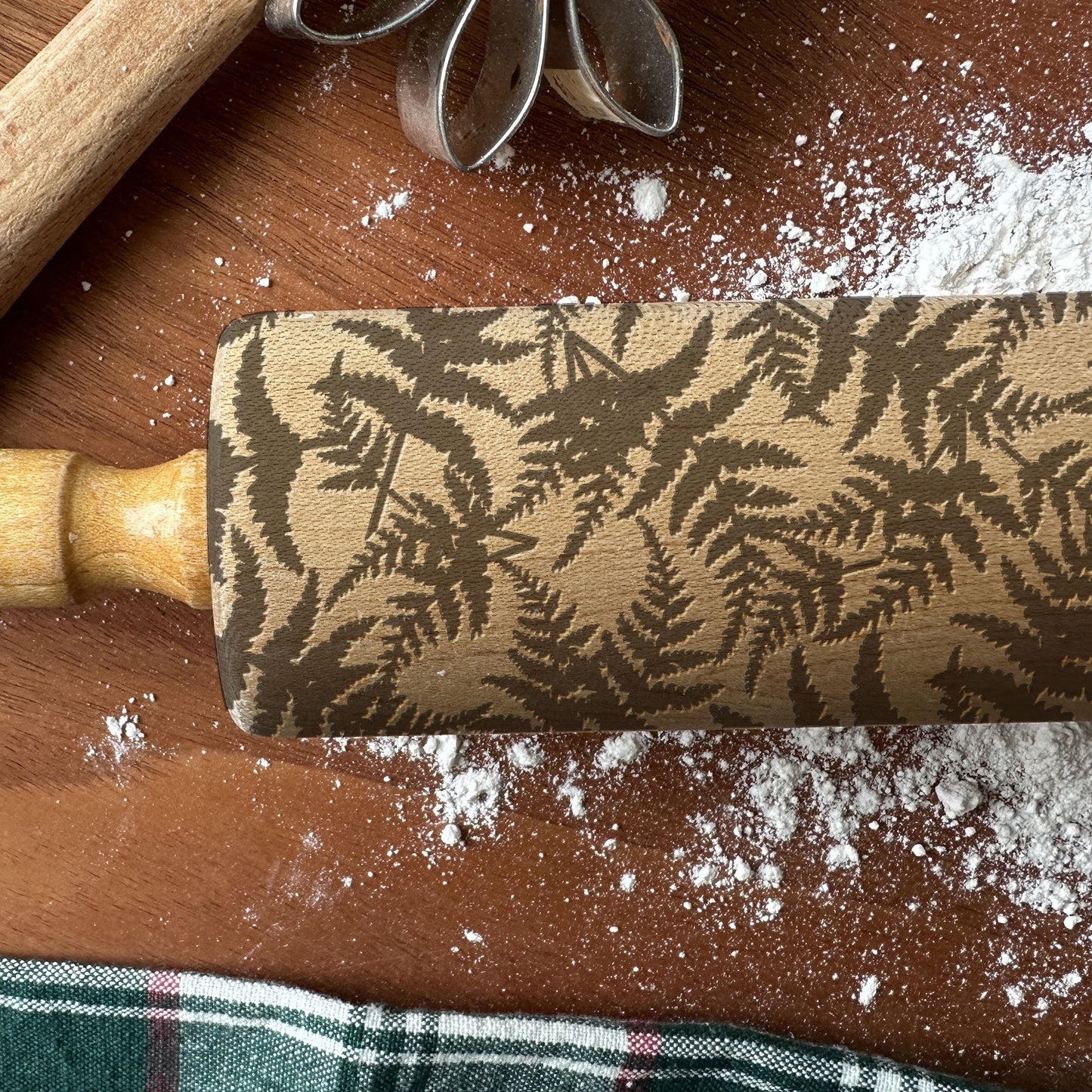 Wooden rolling pin with leaf pattern on a wooden surface with flour and cookie cutters.