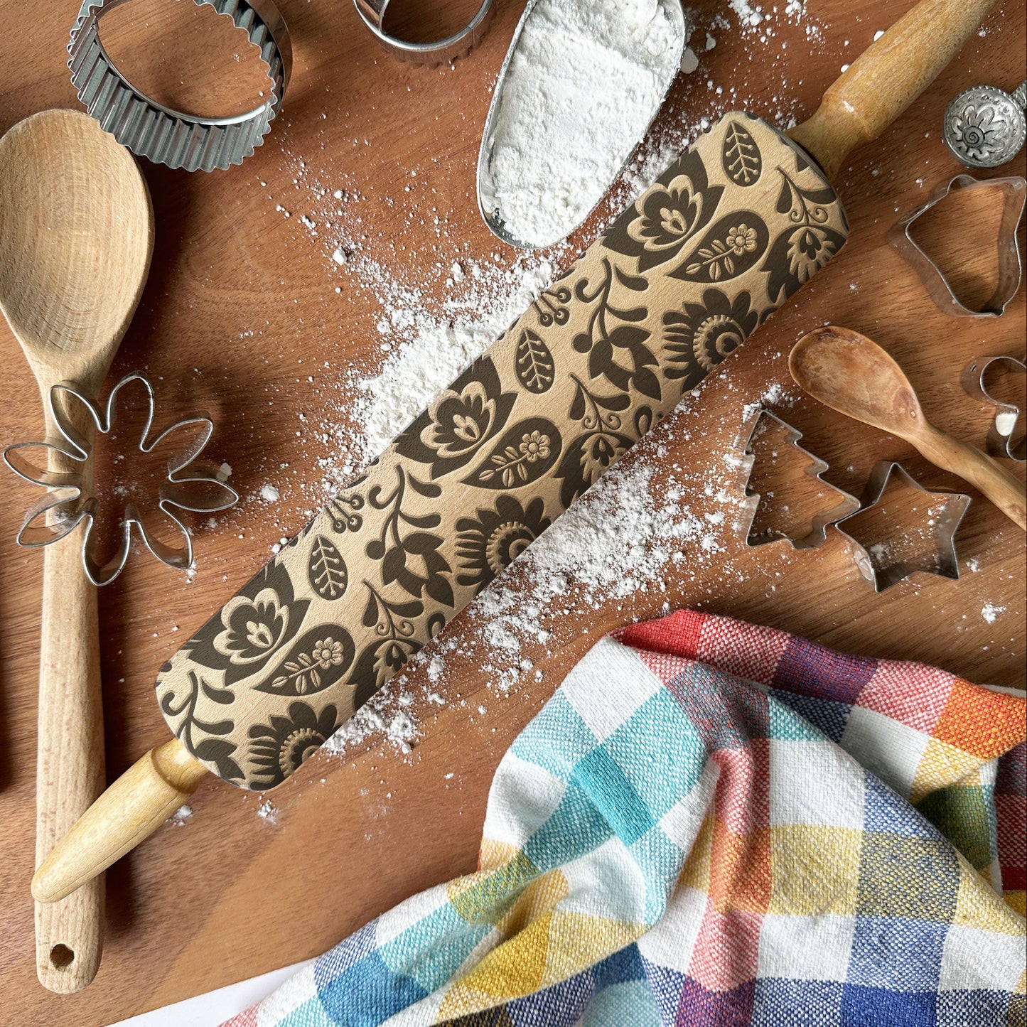 Baking tools including a rolling pin with floral patterns, wooden spoon, whisk, and cookie cutters on a wooden surface with flour.