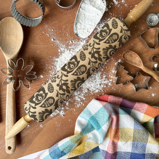 Baking tools including a rolling pin with floral patterns, wooden spoon, whisk, and cookie cutters on a wooden surface with flour.