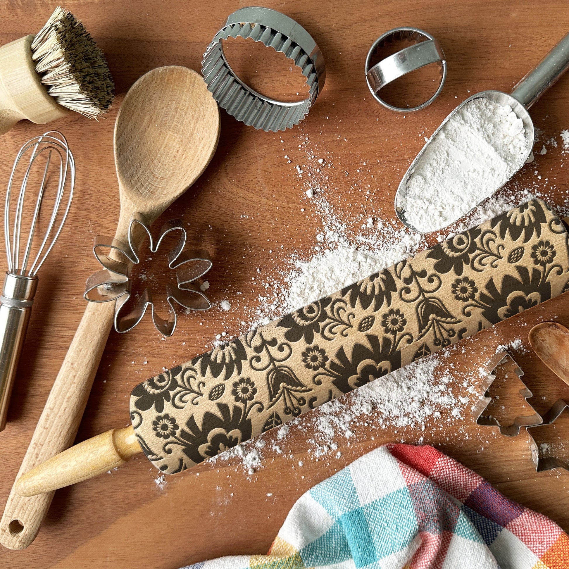 Baking tools including a rolling pin with decorative patterns, wooden spoon, whisk, and cookie cutters on a wooden surface.