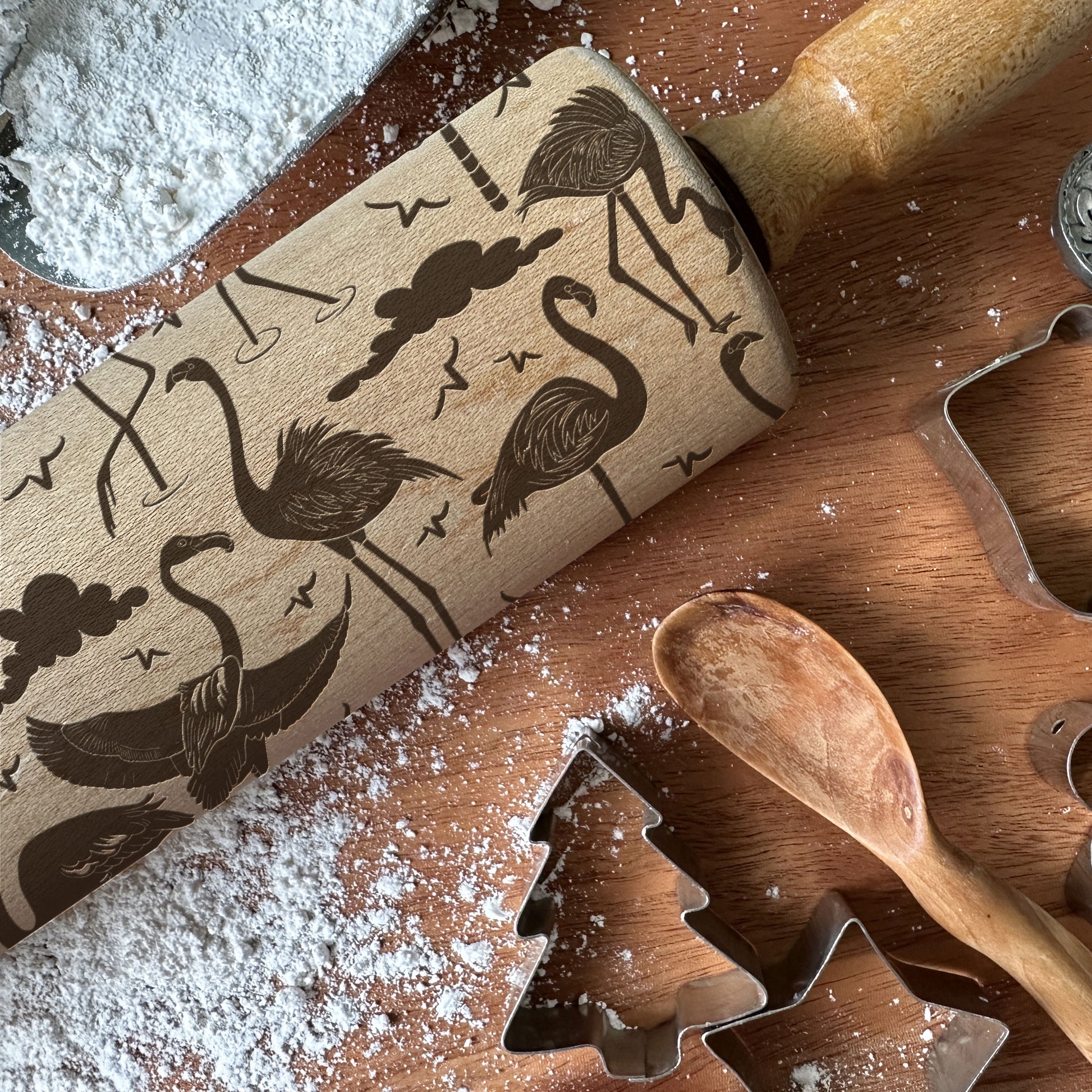 Wooden rolling pin with flamingo design on a wooden surface with flour and cookie cutters.