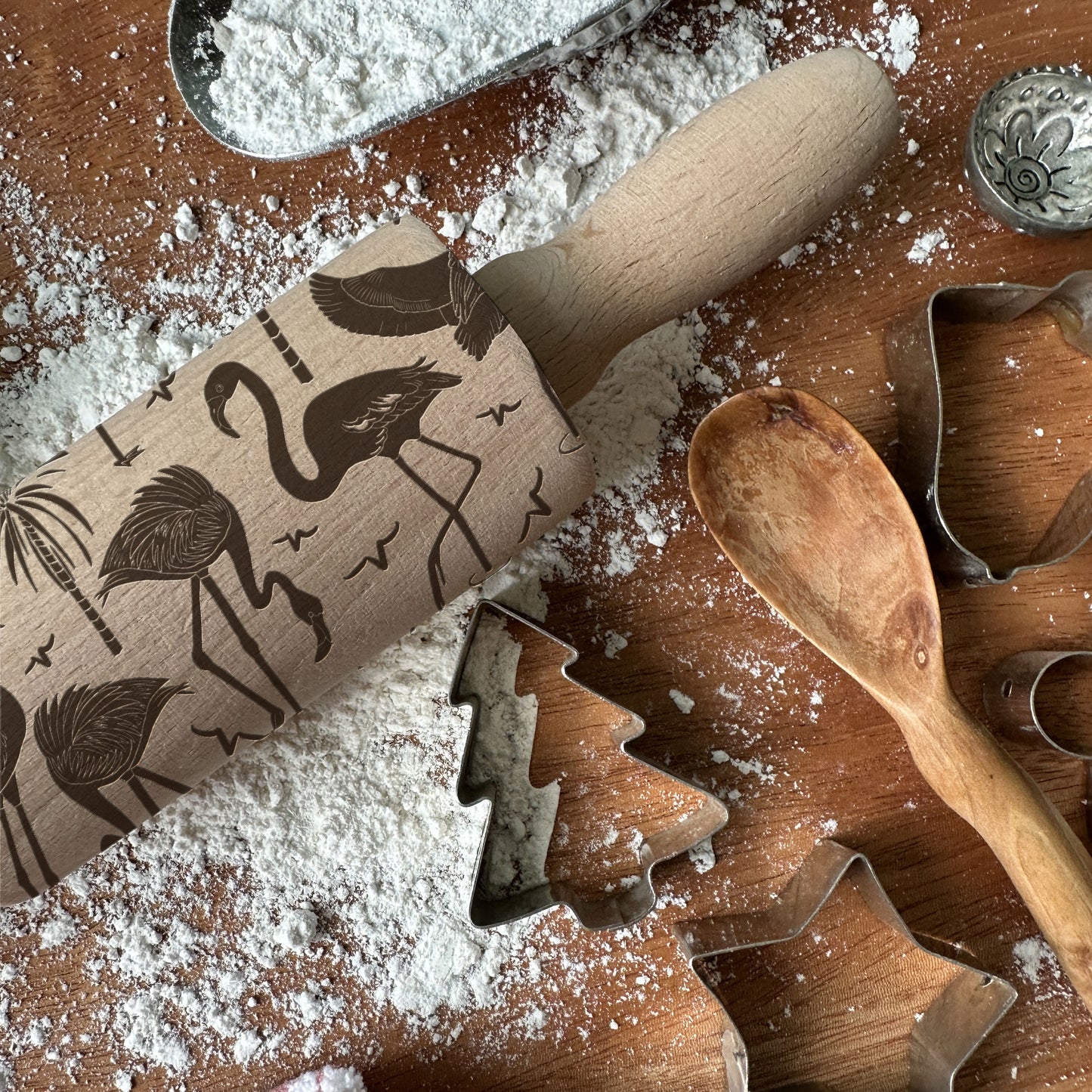 Wooden rolling pin with embossed designs on a flour-dusted surface with cookie cutters and a wooden spoon.