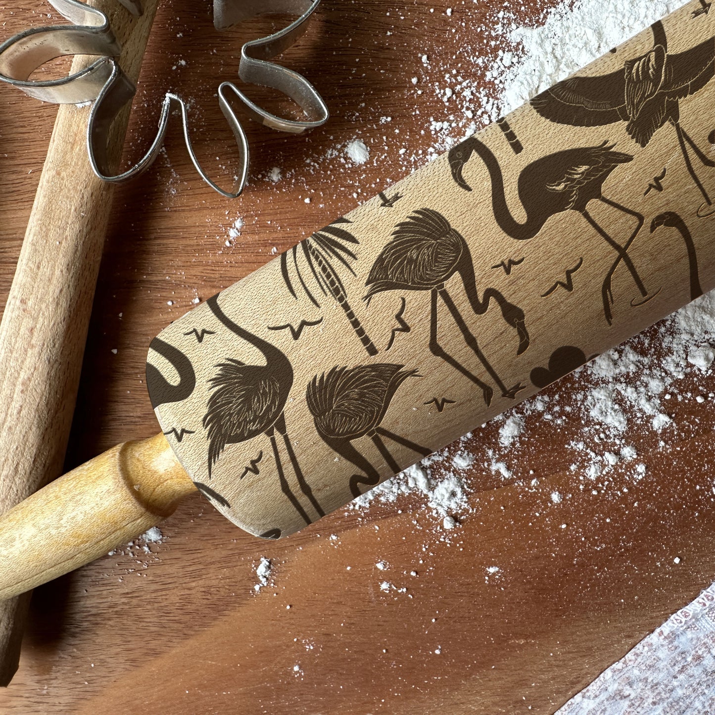 Rolling pin with flamingo pattern on a wooden surface with flour and cookie cutters.