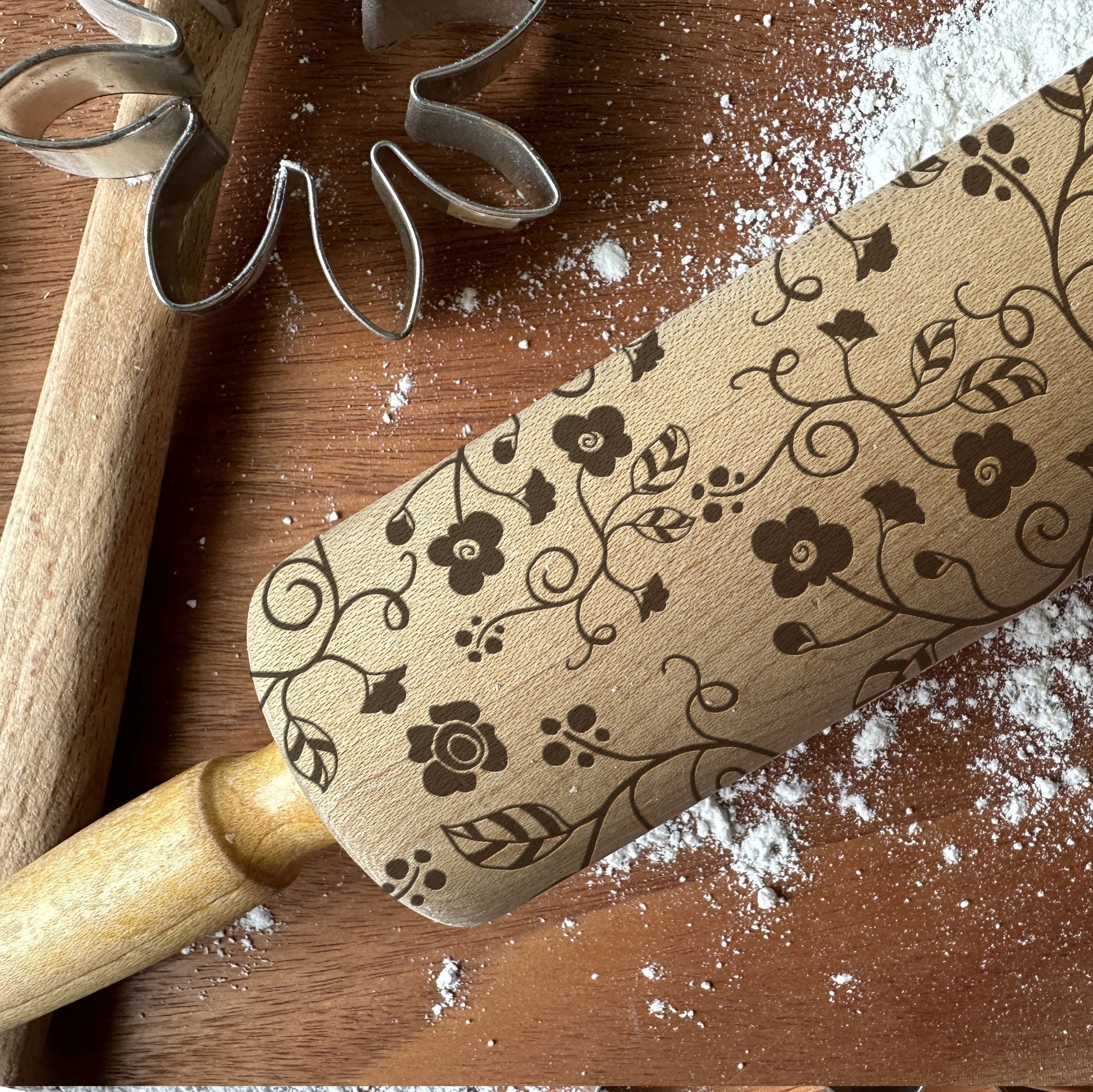 Decorative rolling pin with floral patterns on a wooden surface with flour and cookie cutters.