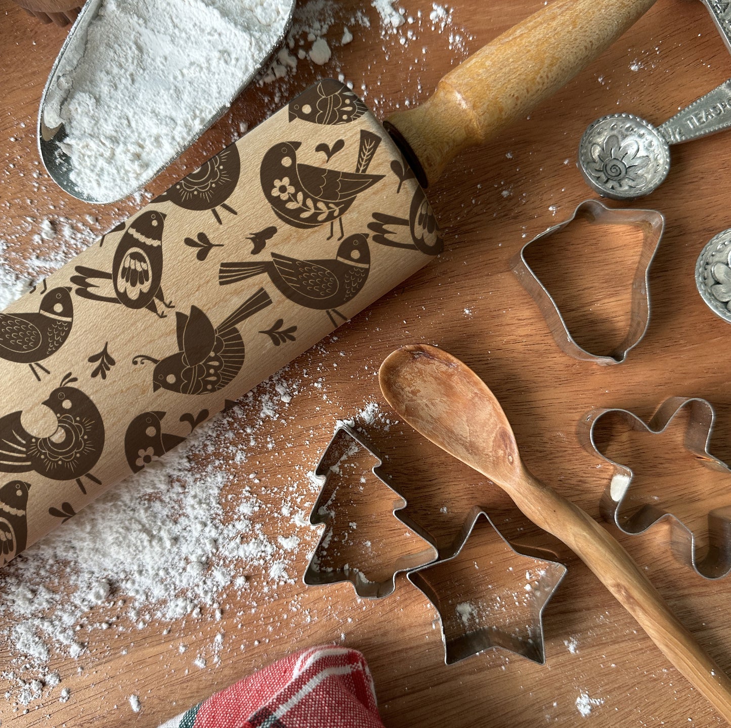 Wooden rolling pin with bird pattern and cookie cutters on a wooden surface with flour.