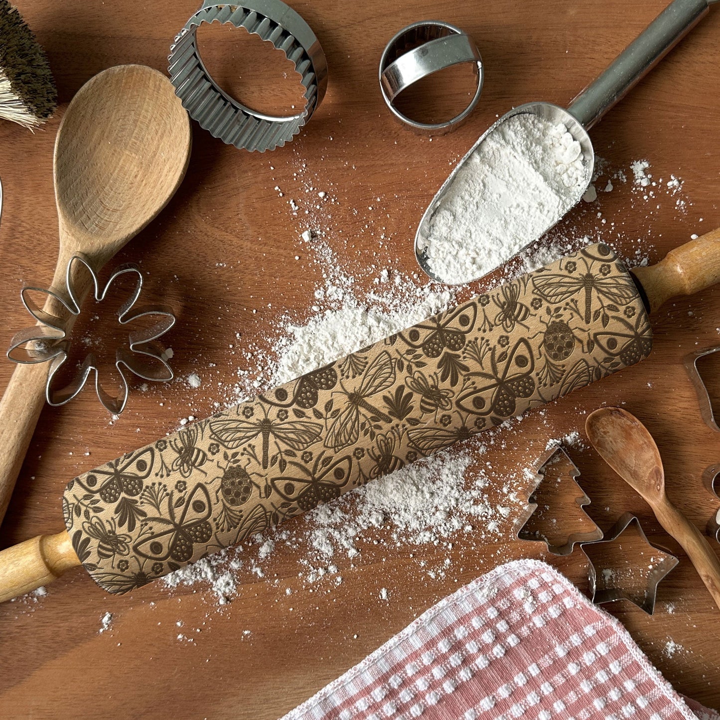 Baking tools including a wooden rolling pin with decorative patterns, cookie cutters, and flour on a wooden surface.