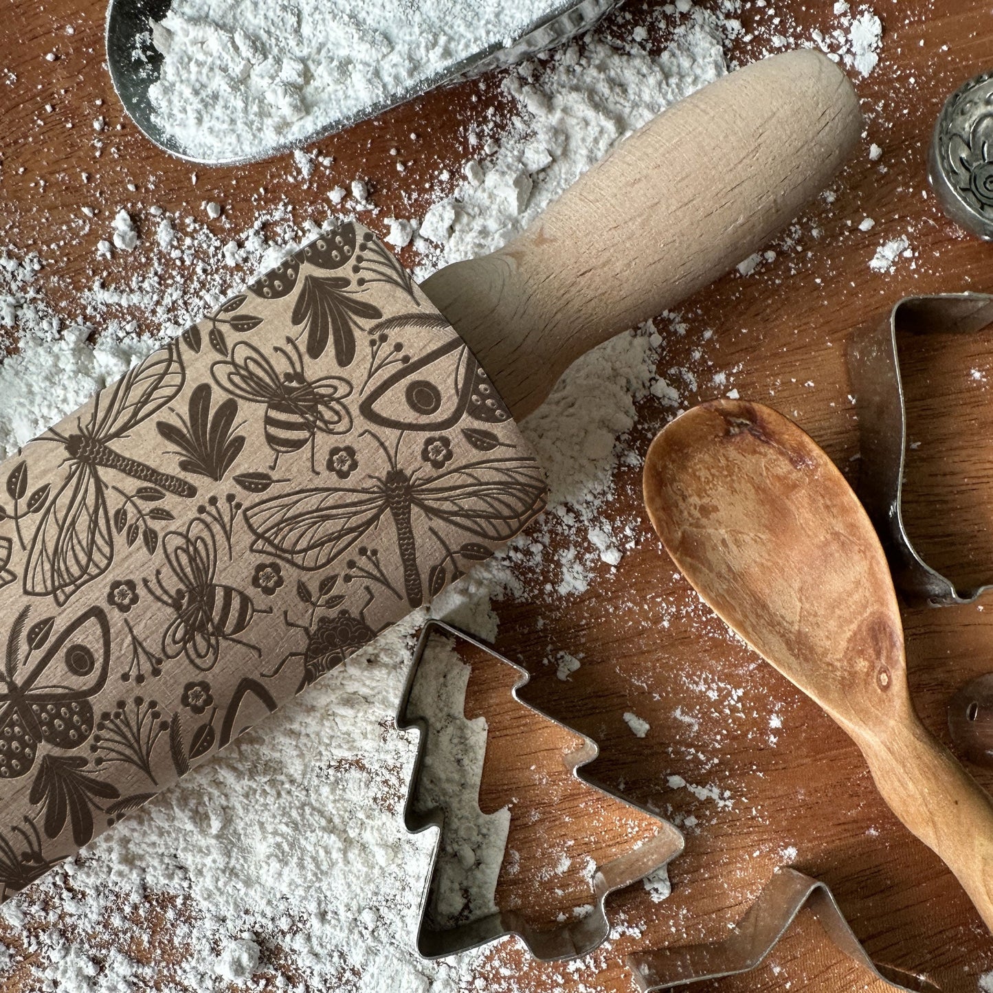 Wooden rolling pin with floral design, wooden spoon, and cookie cutters on a flour-dusted surface.