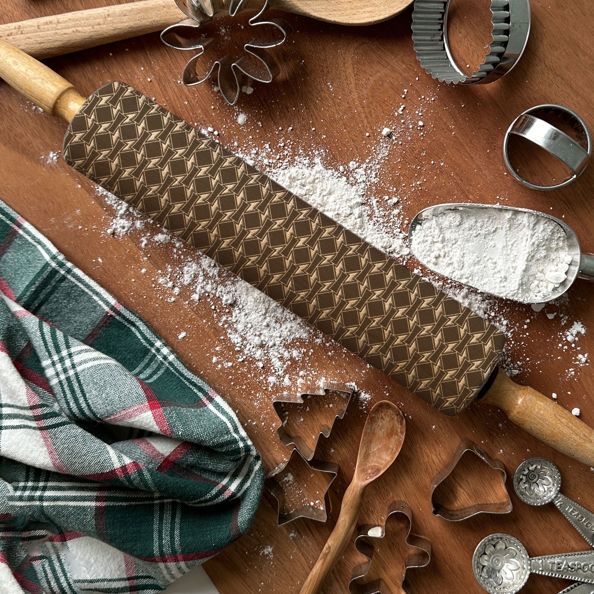Baking tools including a patterned rolling pin, wooden spoons, and metal cookie cutters on a wooden surface with flour.