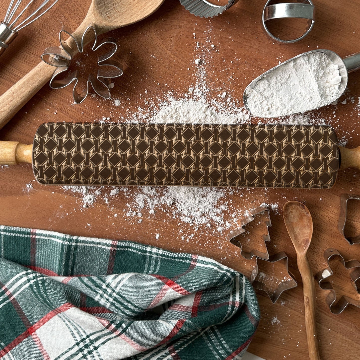 Wooden rolling pin with pattern, flour, plaid cloth, and baking tools on a wooden surface.
