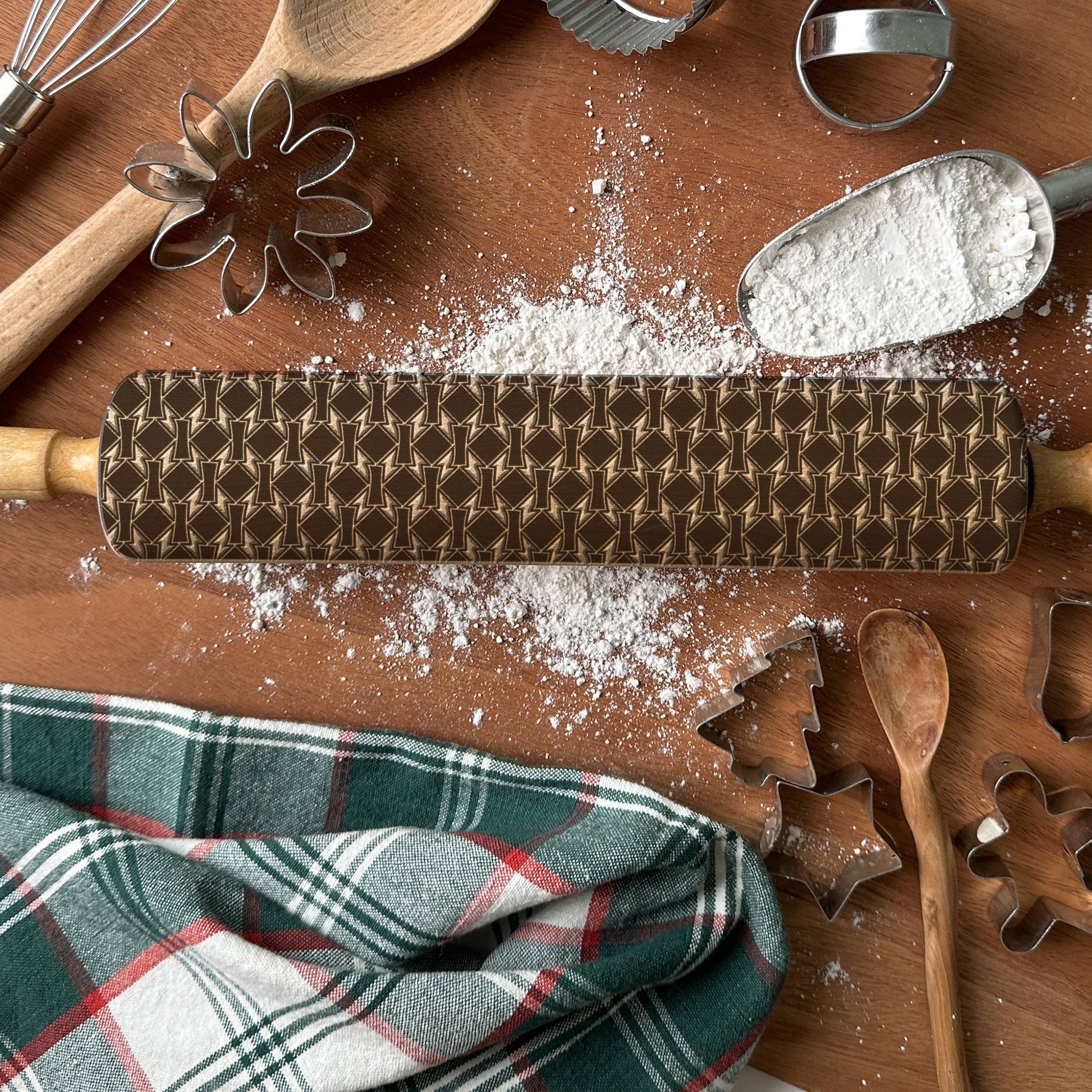 Wooden rolling pin with pattern, flour, plaid cloth, and baking tools on a wooden surface.