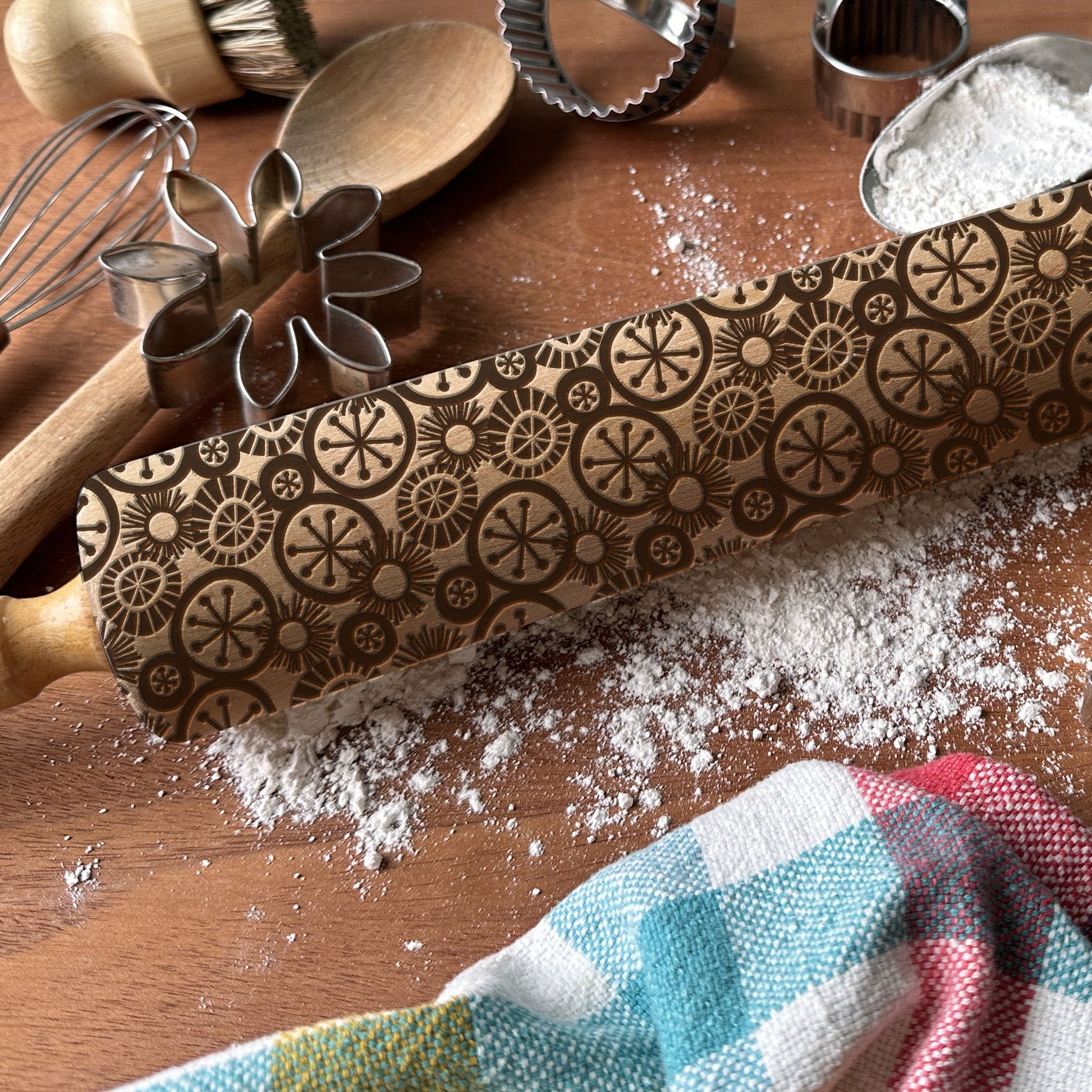 Decorative rolling pin with embossed patterns on a wooden surface with flour and kitchen tools.