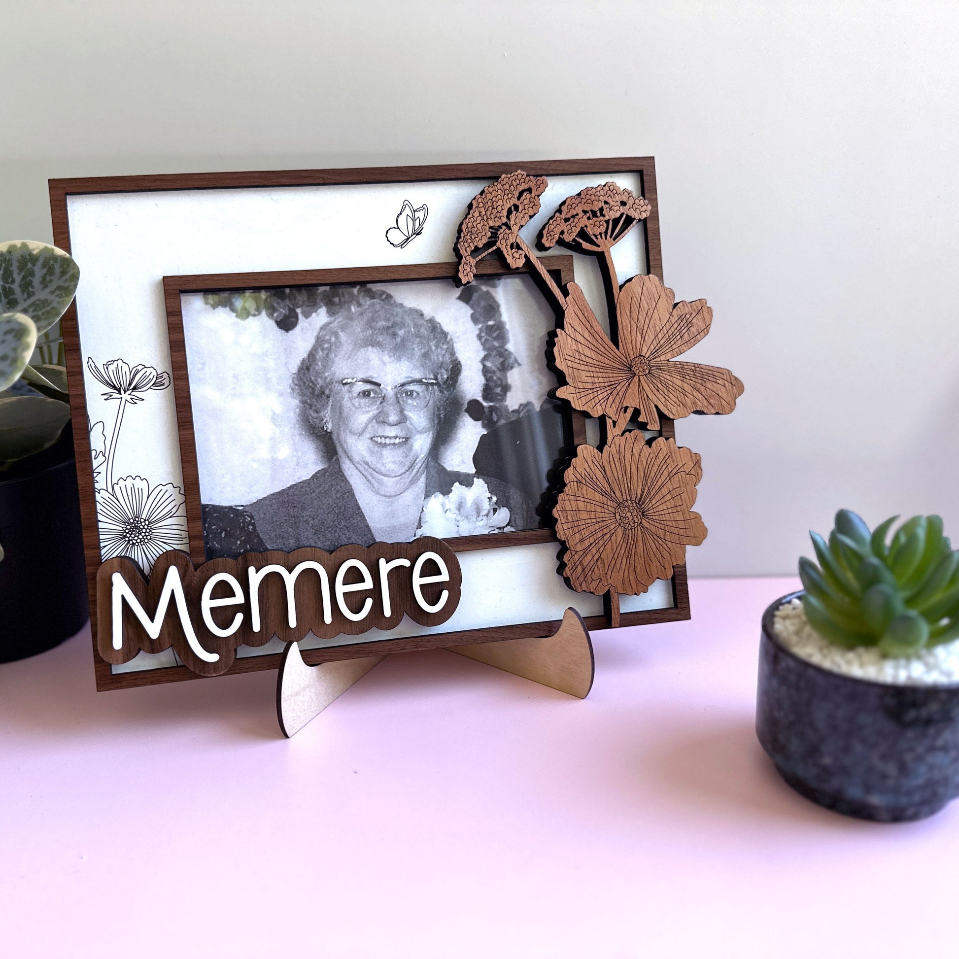 Decorative photo frame with a black and white photo of an elderly woman, wooden butterflies, and the word 'Memere' on a light pink surface.