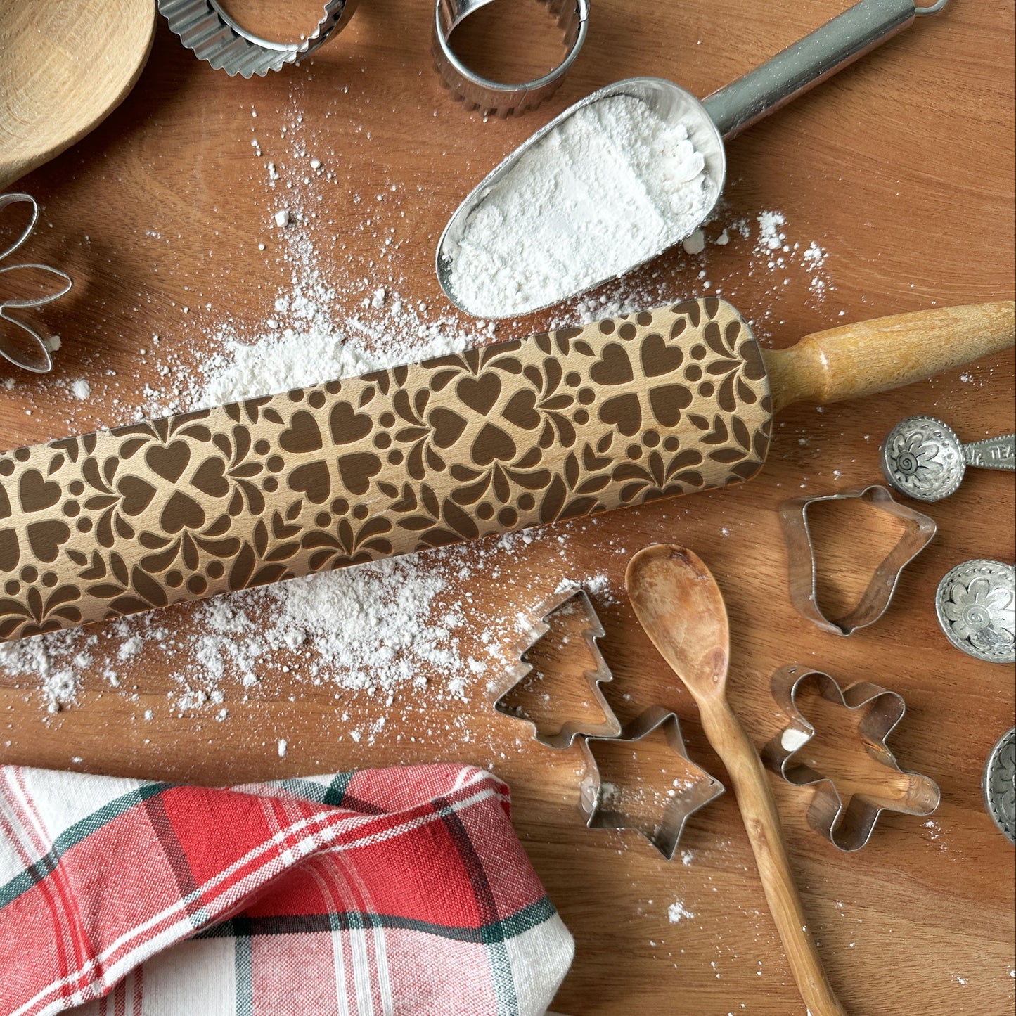 Wooden rolling pin with embossed patterns on a wooden surface with flour, cookie cutters, and a plaid cloth.