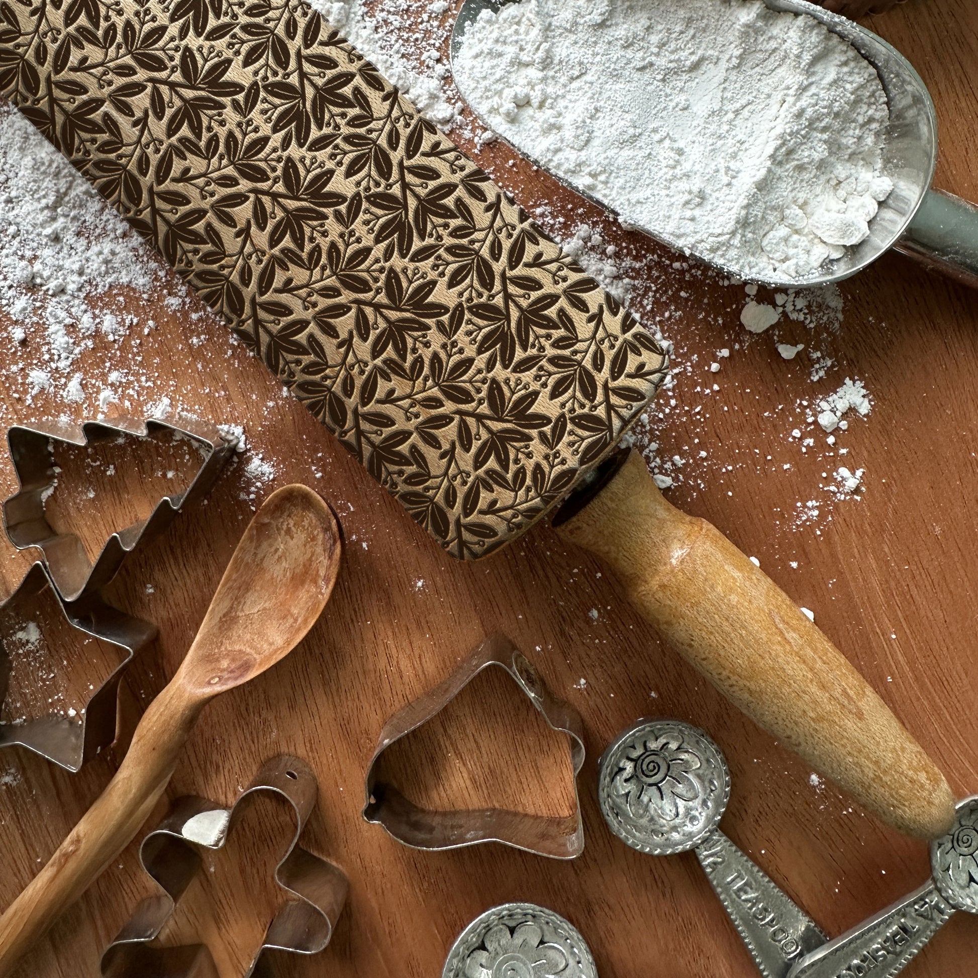Wooden rolling pin with leaf pattern, flour, wooden spoon, and metal cookie cutters on a wooden surface.