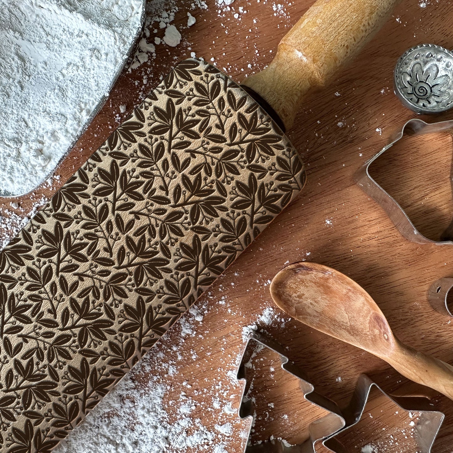 Wooden rolling pin with leaf pattern and cookie cutters on a wooden surface with flour.