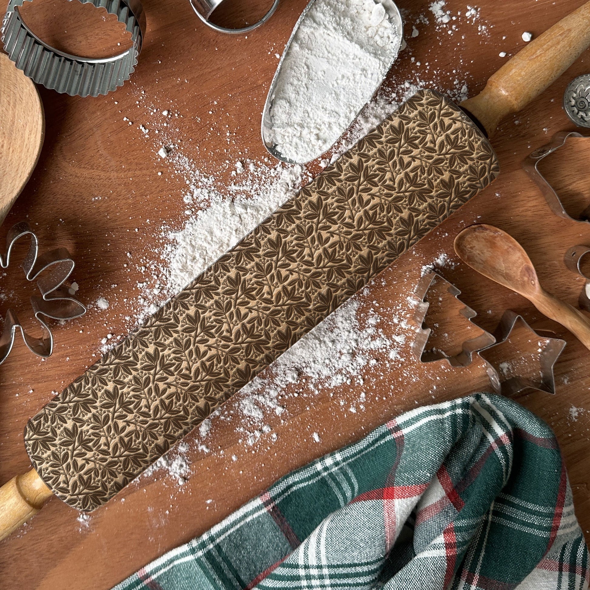 Baking tools including a rolling pin with embossed patterns, cookie cutters, and a plaid towel on a wooden surface.