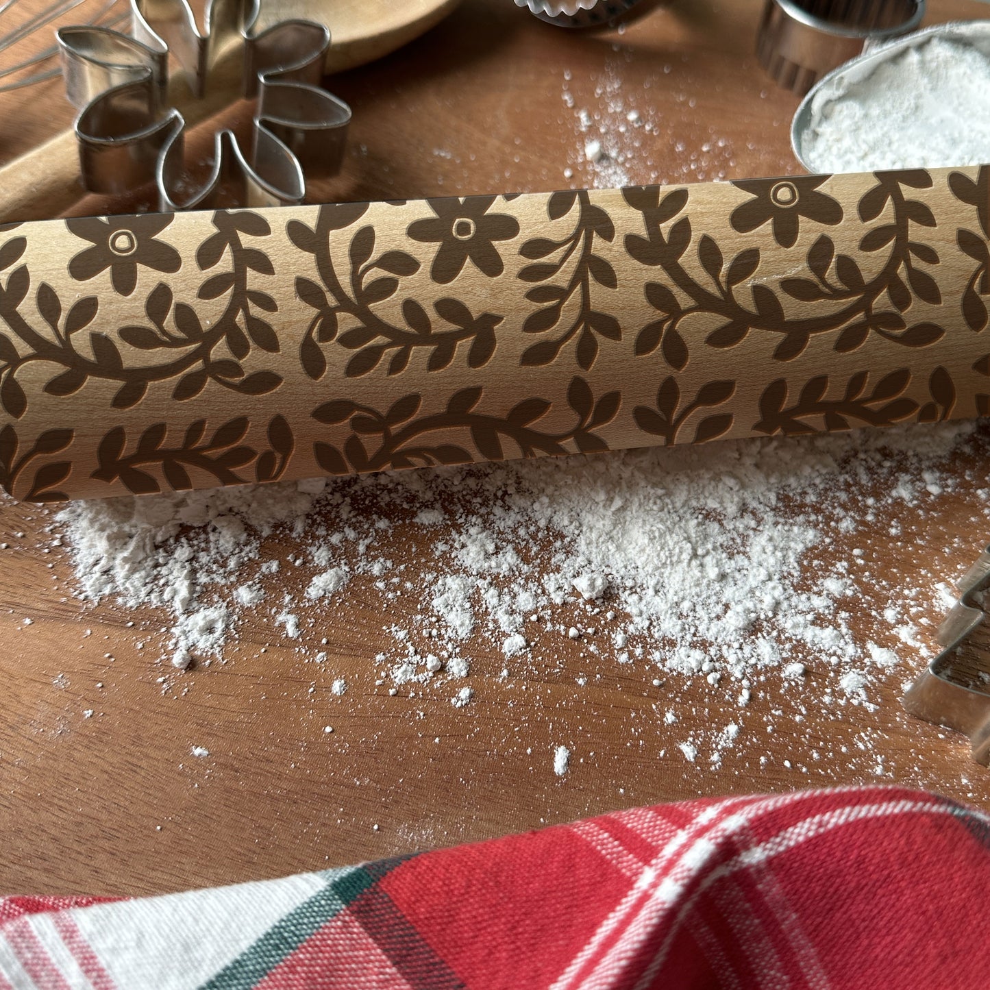 Decorative rolling pin with floral pattern on a wooden surface with flour and cookie cutters.
