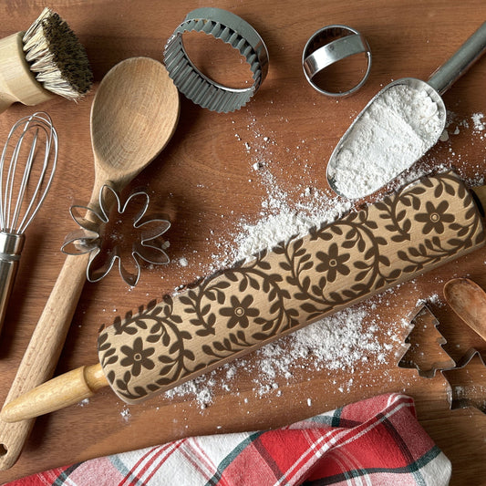 Baking tools including a rolling pin with floral patterns, wooden spoon, whisk, and cookie cutters on a wooden surface.