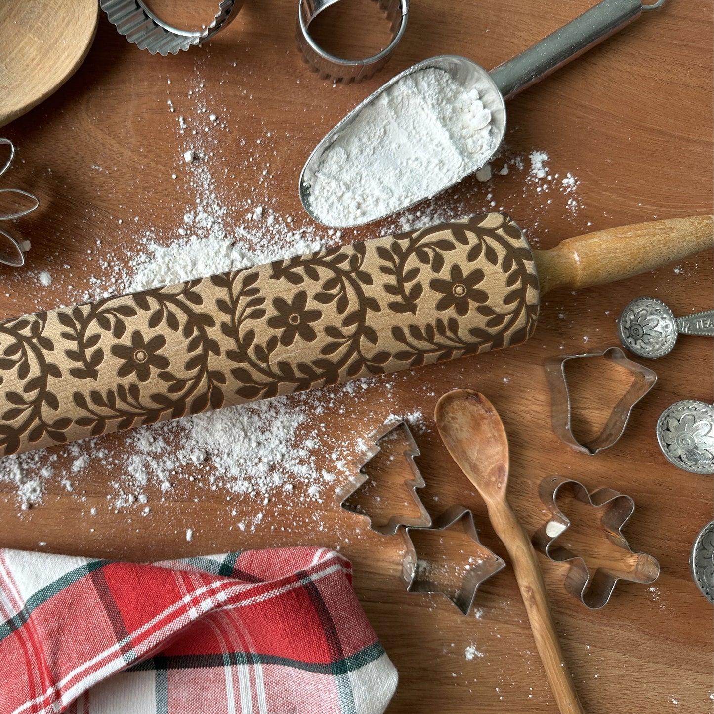 Decorative rolling pin with floral patterns on a wooden surface with flour, cookie cutters, and a plaid cloth.