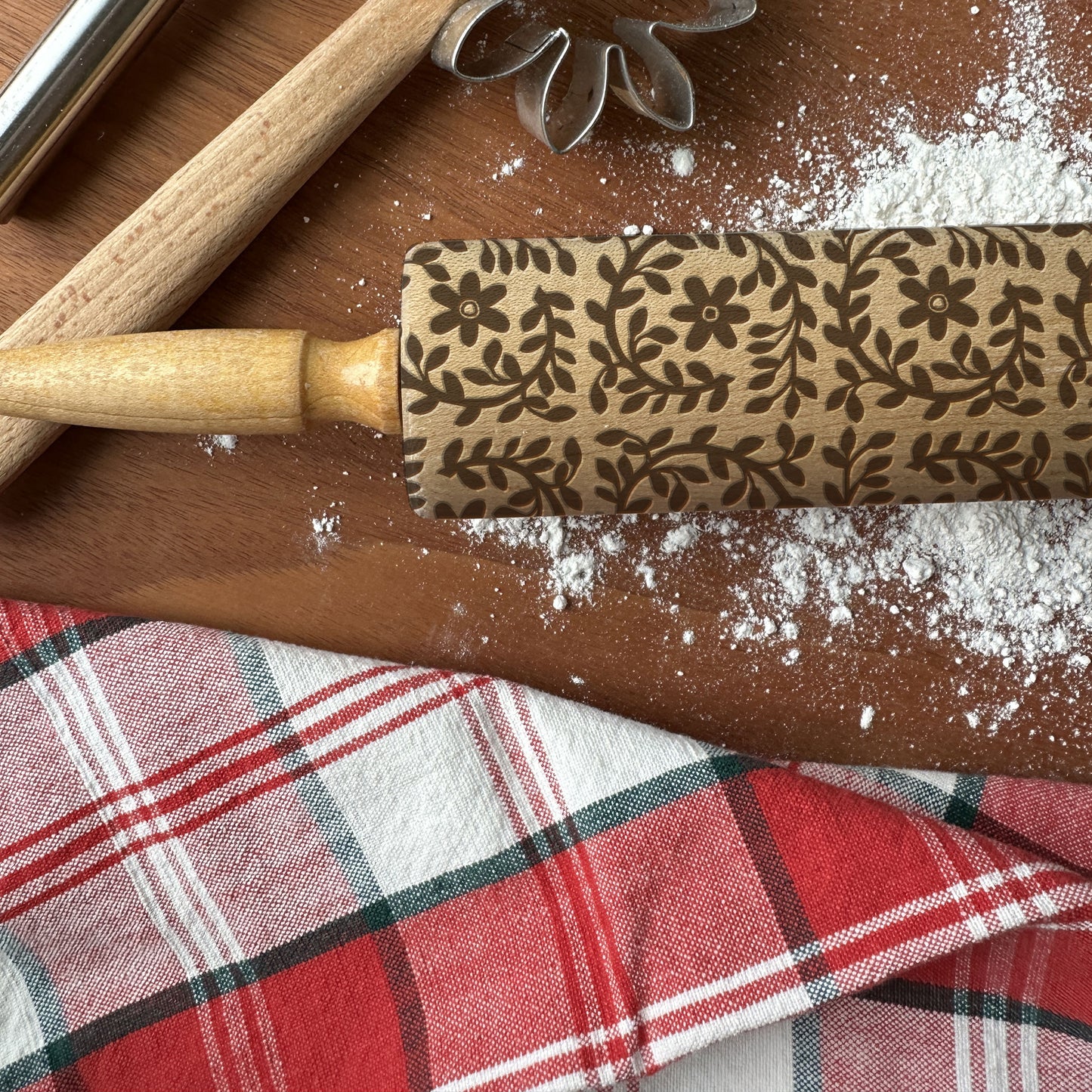 Decorative rolling pin with floral pattern on a wooden surface with flour and plaid fabric.