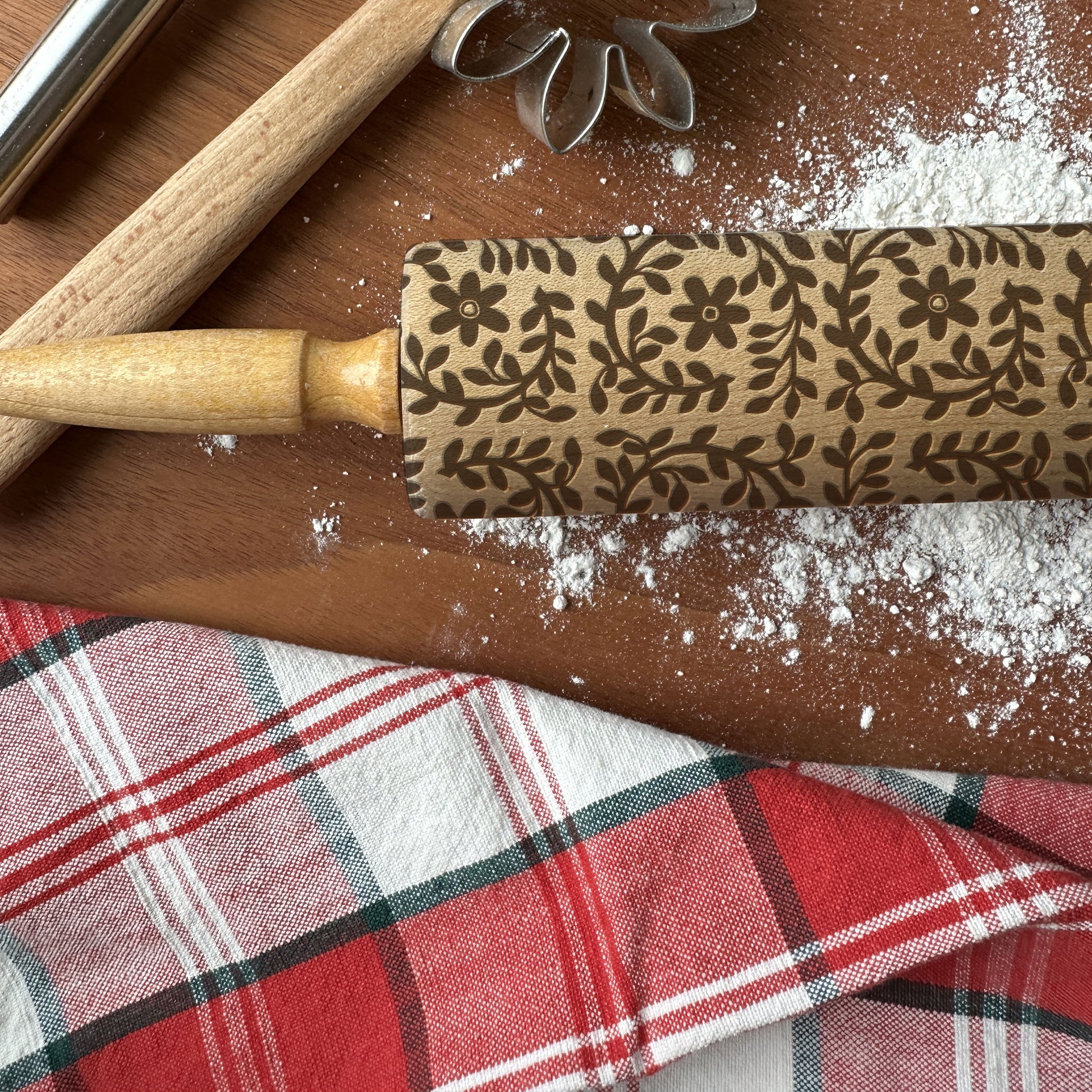 Decorative rolling pin with floral pattern on a wooden surface with flour and plaid fabric.
