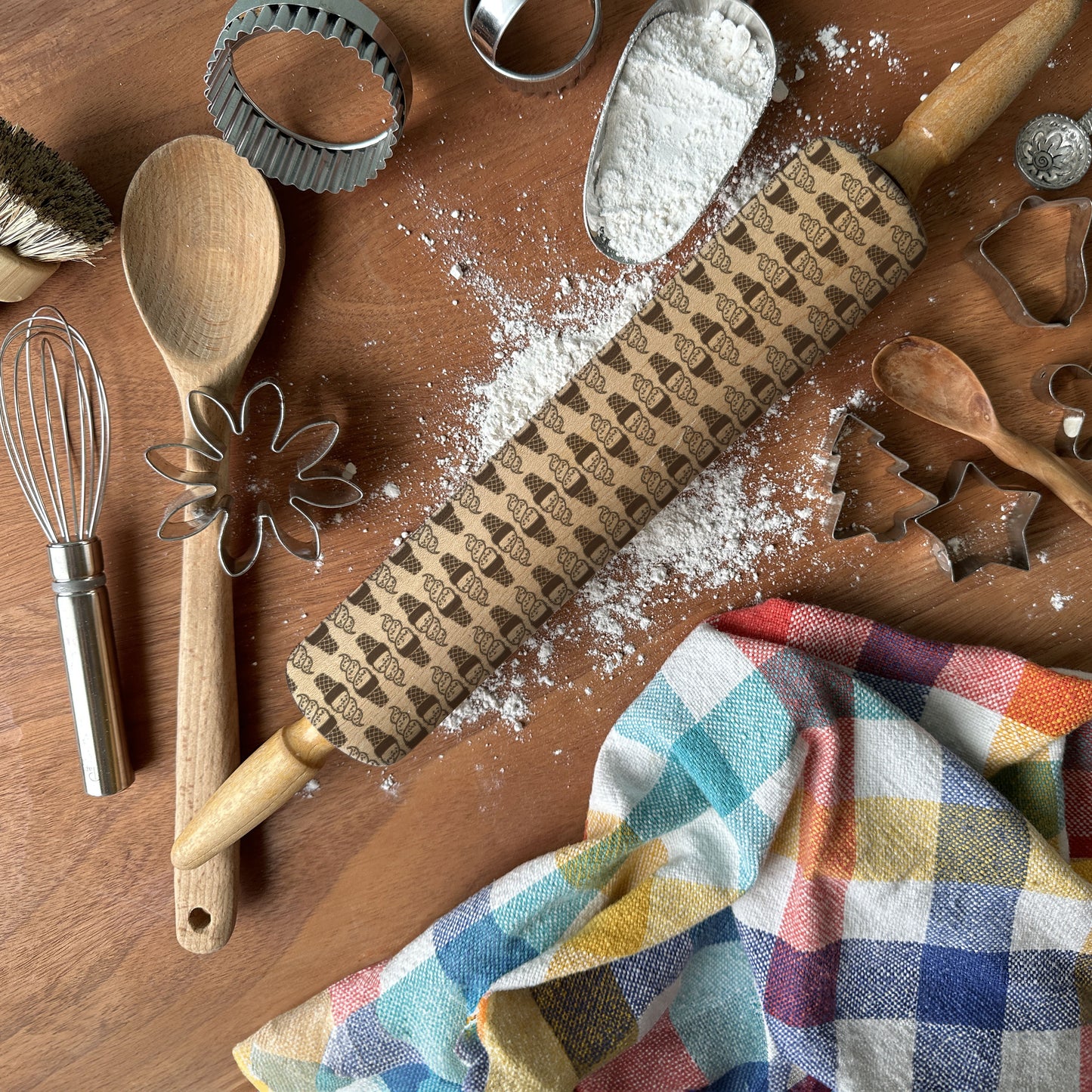 Baking tools including a rolling pin with embossed designs, wooden spoon, whisk, and cookie cutters on a wooden surface with flour.