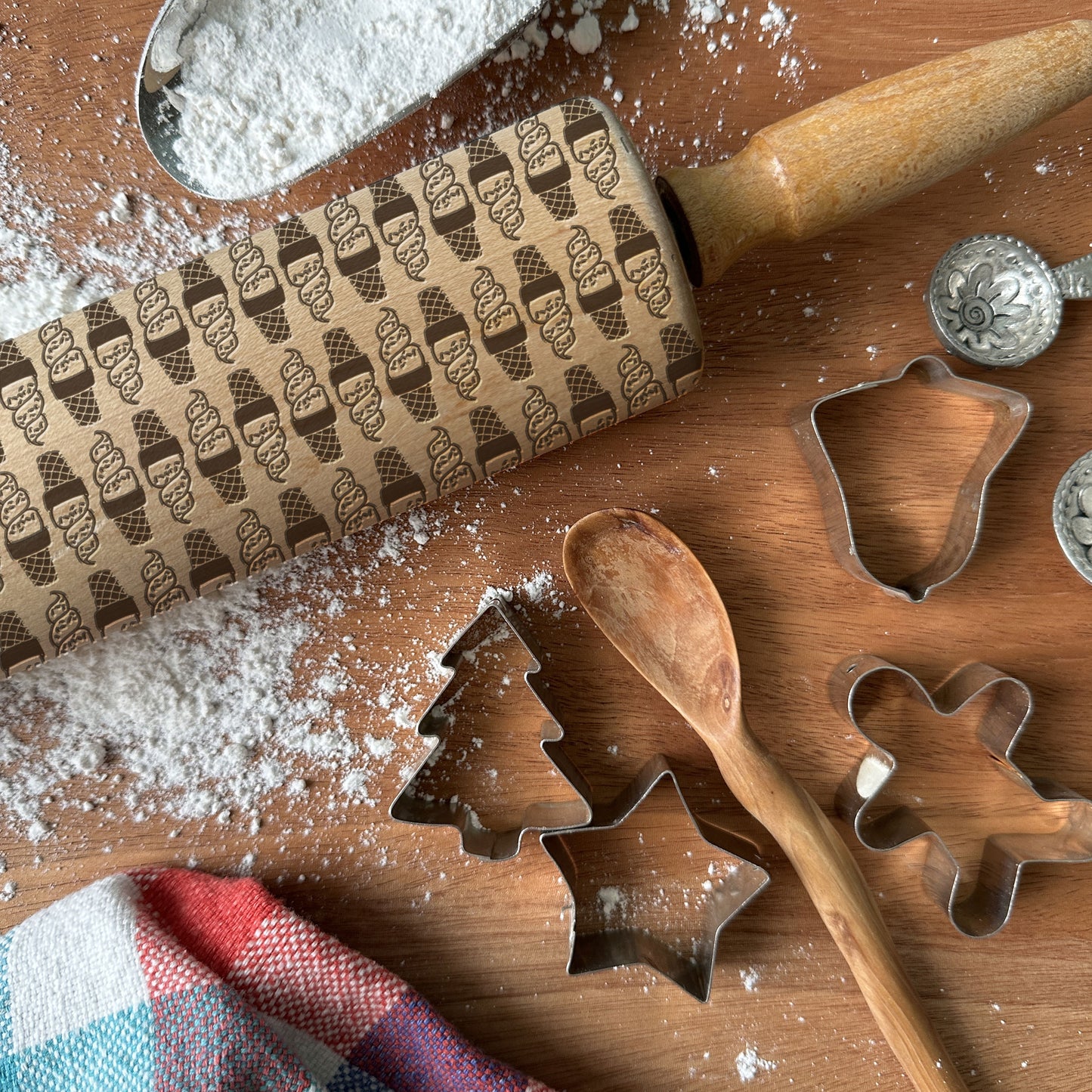 Baking tools including a patterned rolling pin, cookie cutters, and a wooden spoon on a wooden surface with flour.
