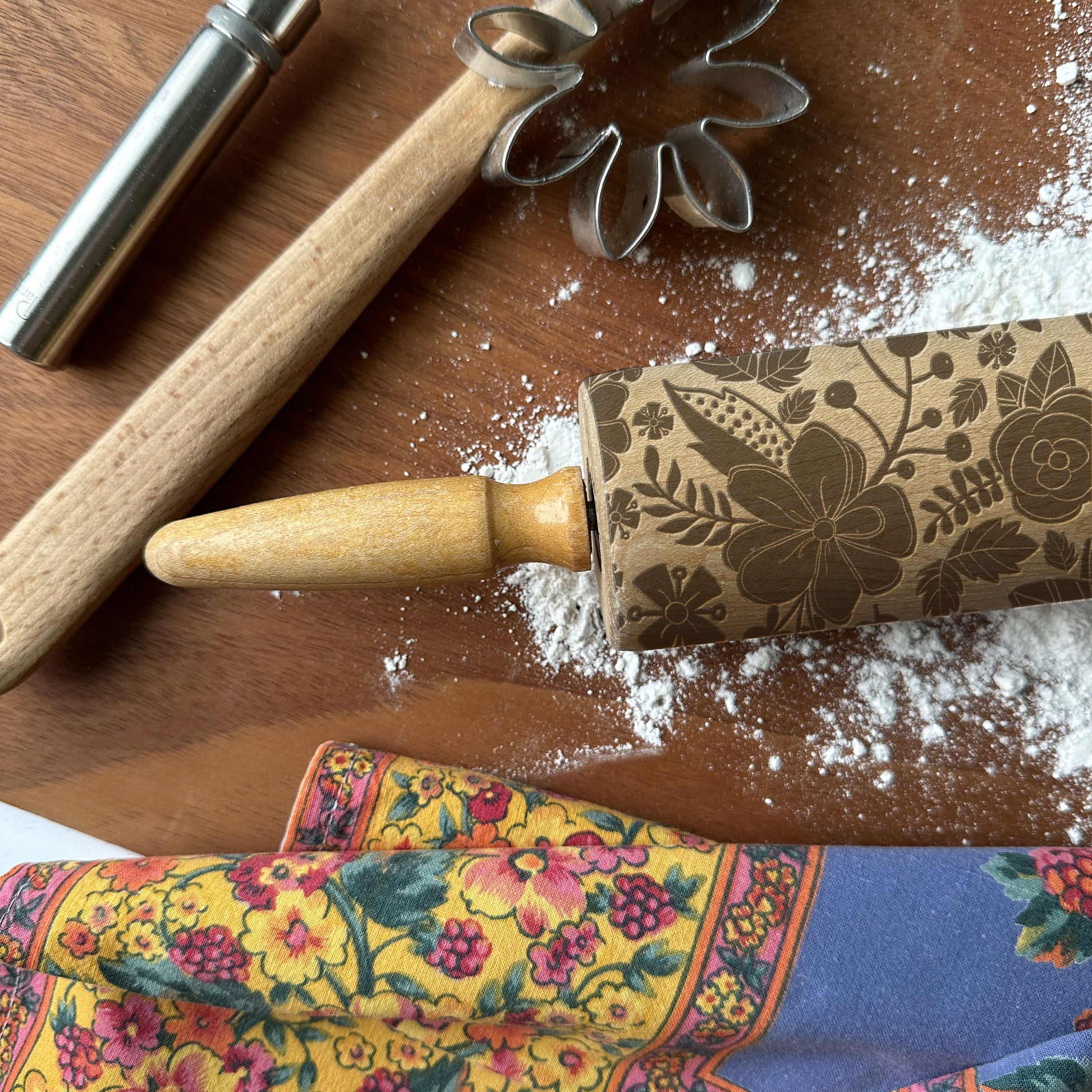 Floral-patterned rolling pin on a wooden surface with flour and cookie cutters.