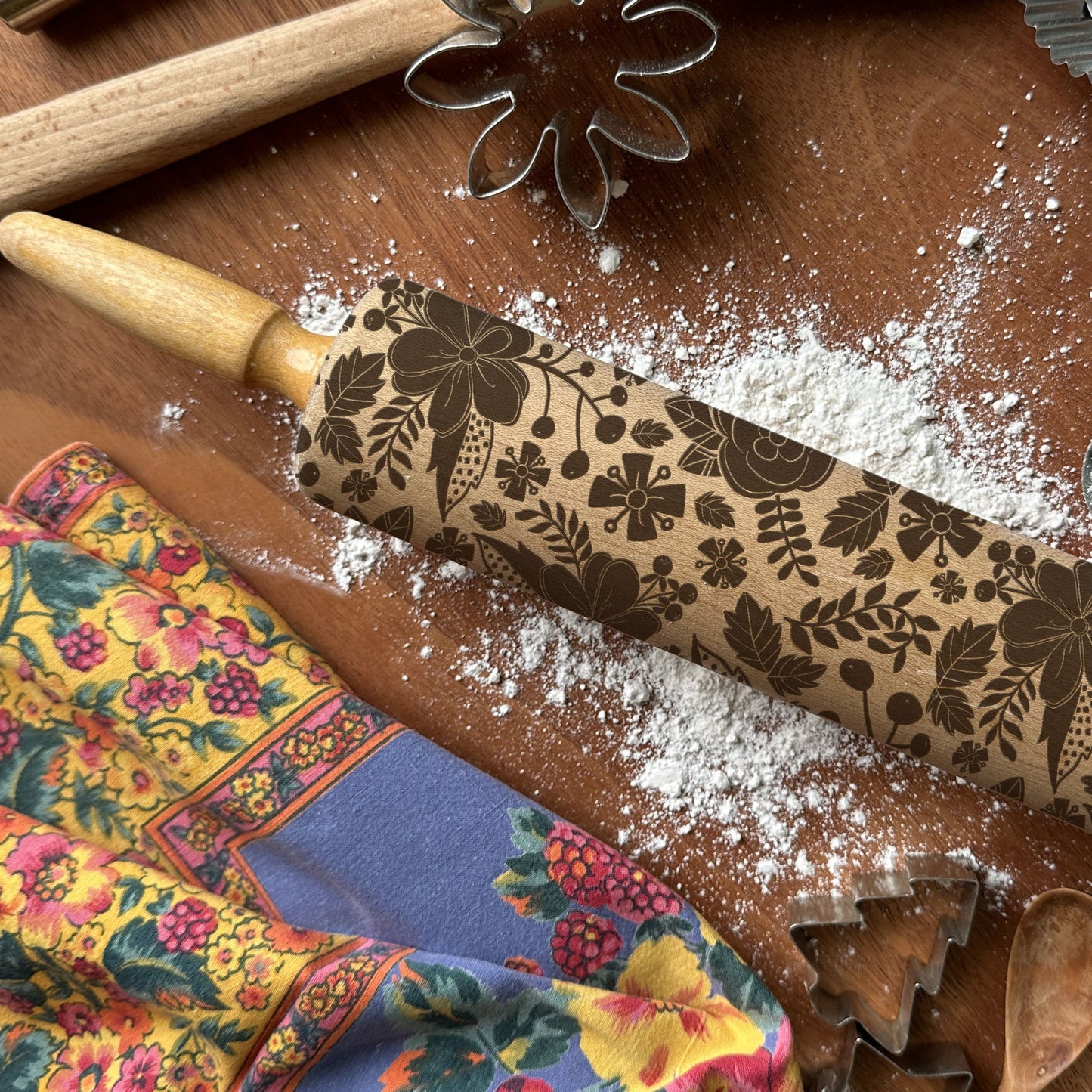 Decorative rolling pin with floral patterns on a wooden surface with flour and cookie cutters.