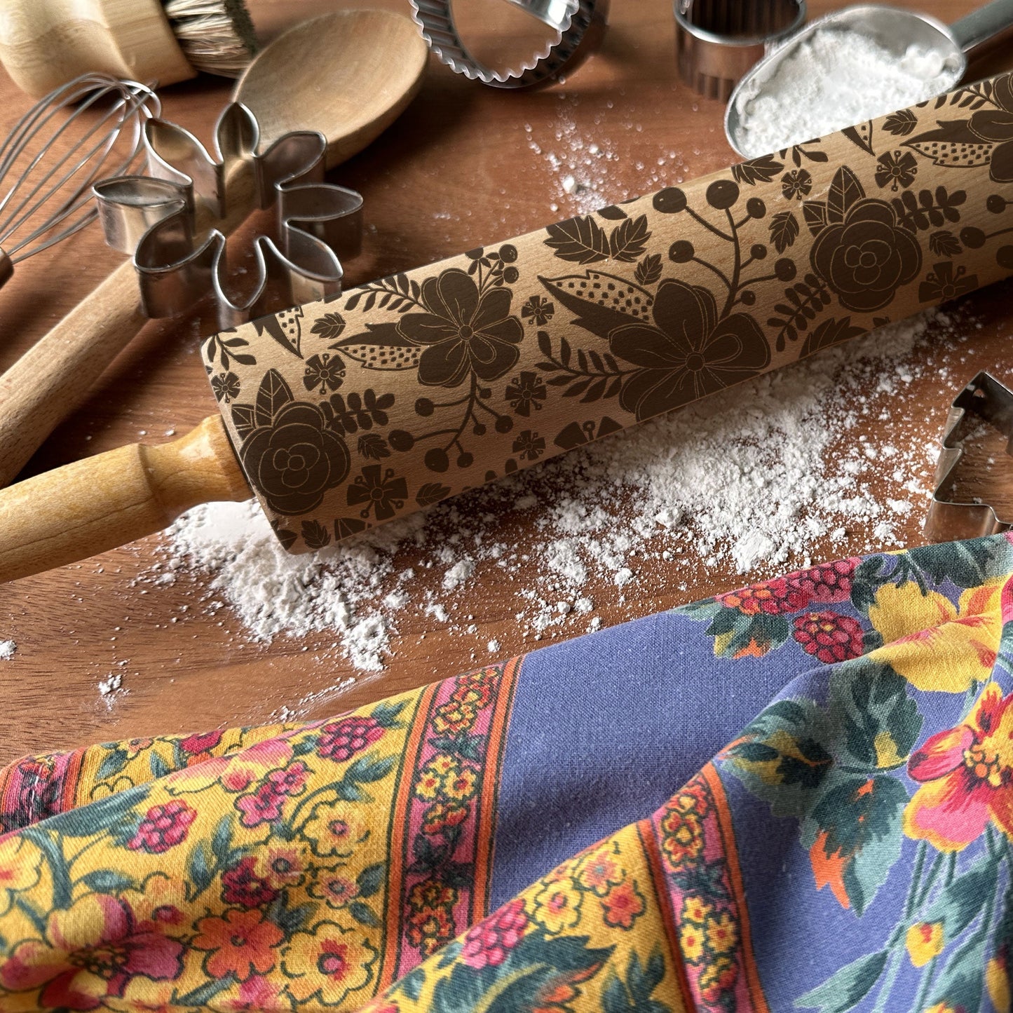 Floral-patterned rolling pin on a wooden surface with kitchen utensils and a colorful towel.