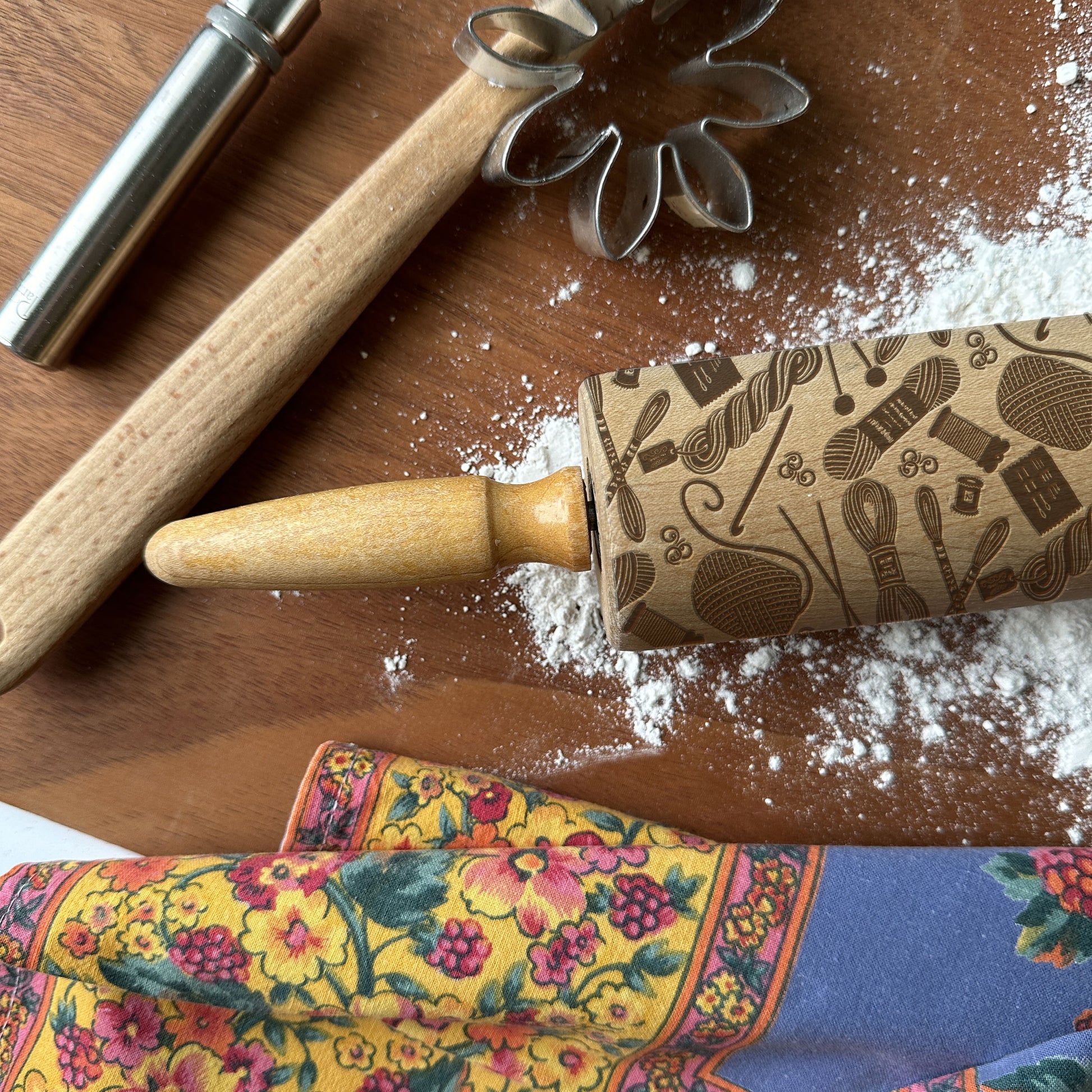 Wooden rolling pin with patterned handle on a wooden surface with flour and cookie cutters.