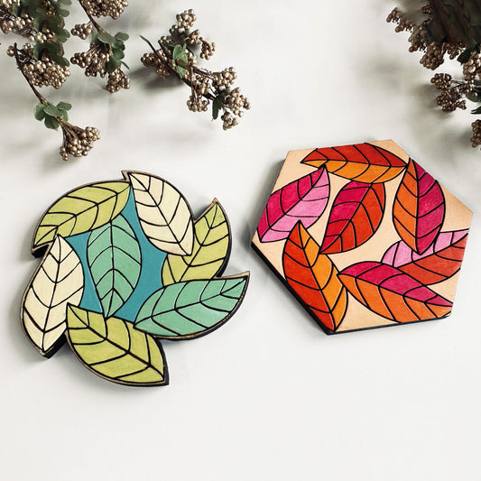 Two colorful leaf-shaped coasters on a light background with dried plants.