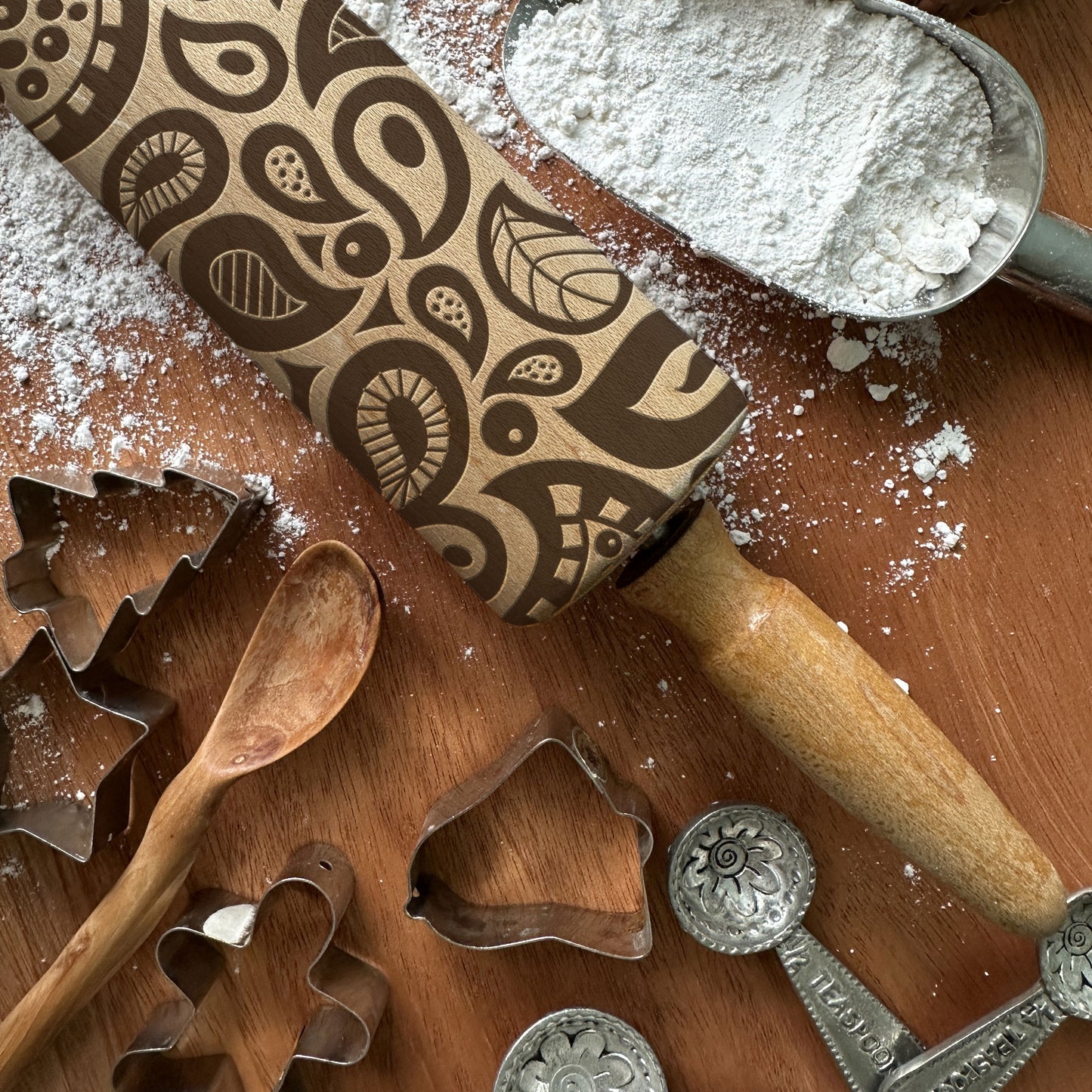 Wooden rolling pin with pattern, flour, wooden spoon, and metal cookie cutters on a wooden surface.
