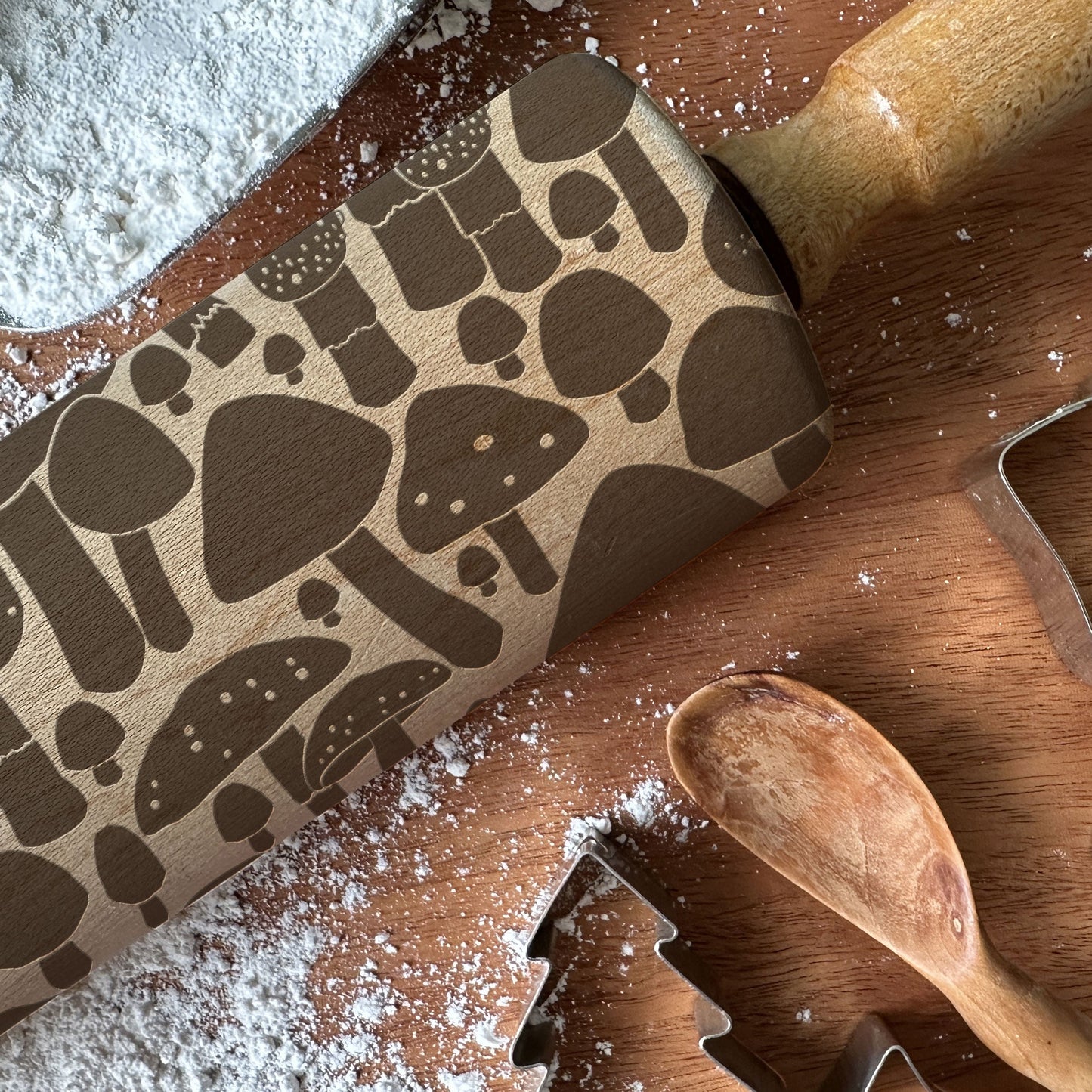 Wooden rolling pin with mushroom pattern on a wooden surface with flour and cookie cutters.