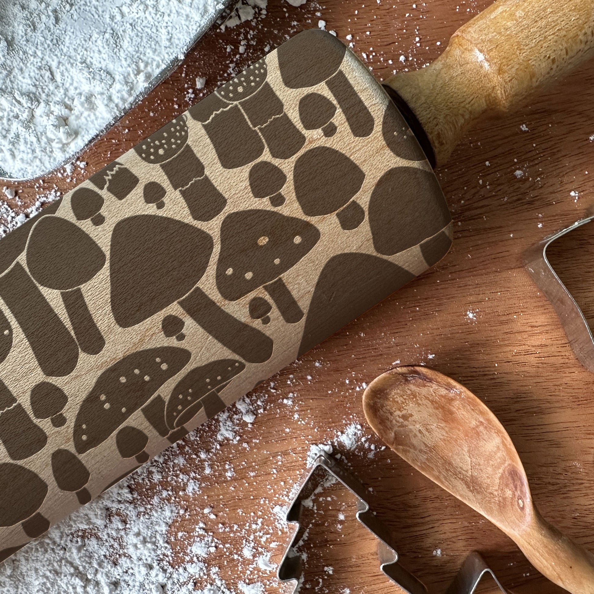 Wooden rolling pin with mushroom pattern on a wooden surface with flour and cookie cutters.
